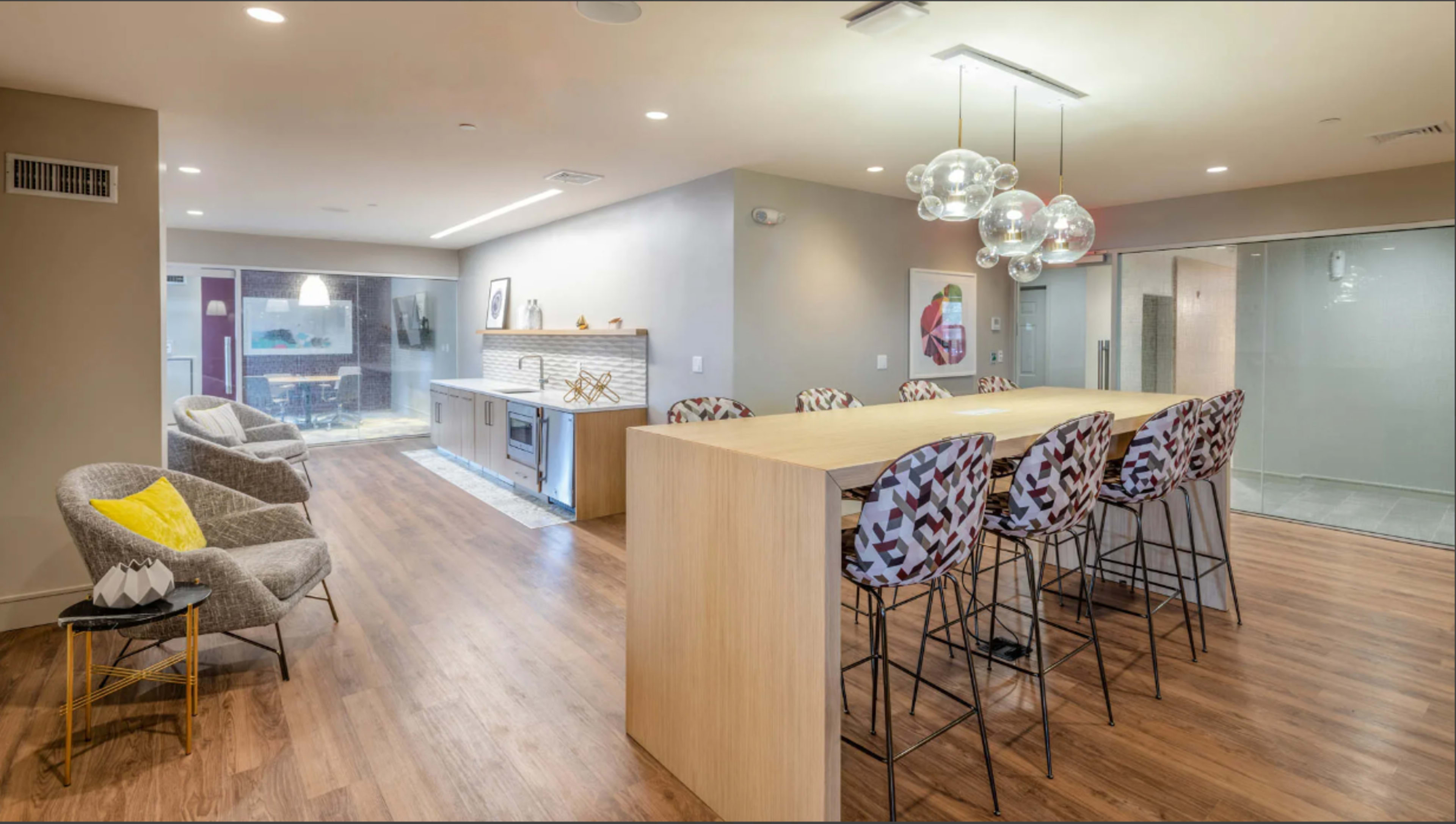 A modern kitchen and dining area with a long wooden bar surrounded by stylish high chairs, and a living area featuring a cozy chair.