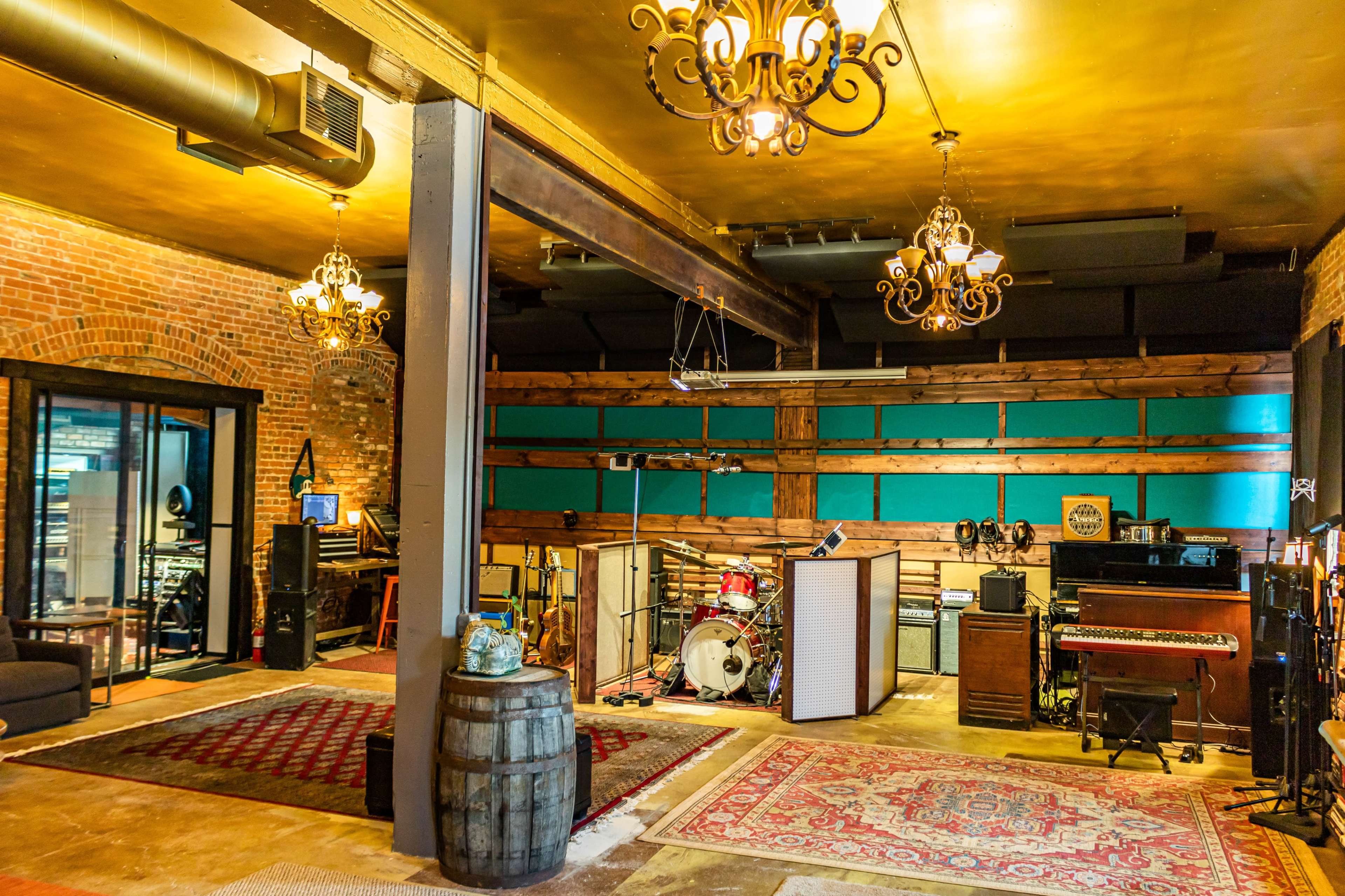 A well-decorated music studio featuring vintage instruments, a drum set, and a large sound booth, all set in a rustic interior with exposed brick walls and chandeliers.