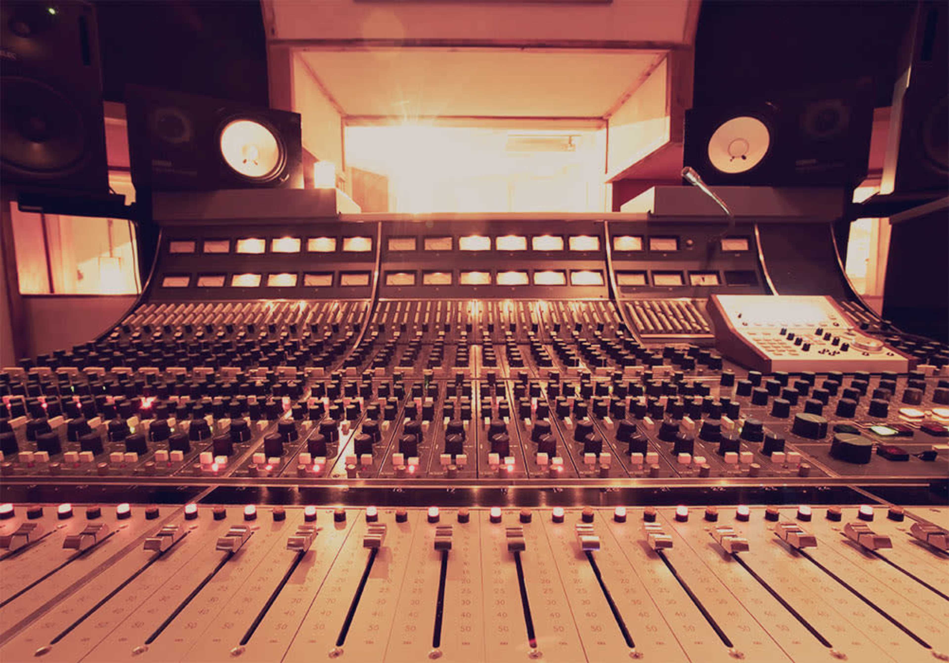 A close-up view of a recording studio mixing console with various knobs, sliders, and speakers in the background.