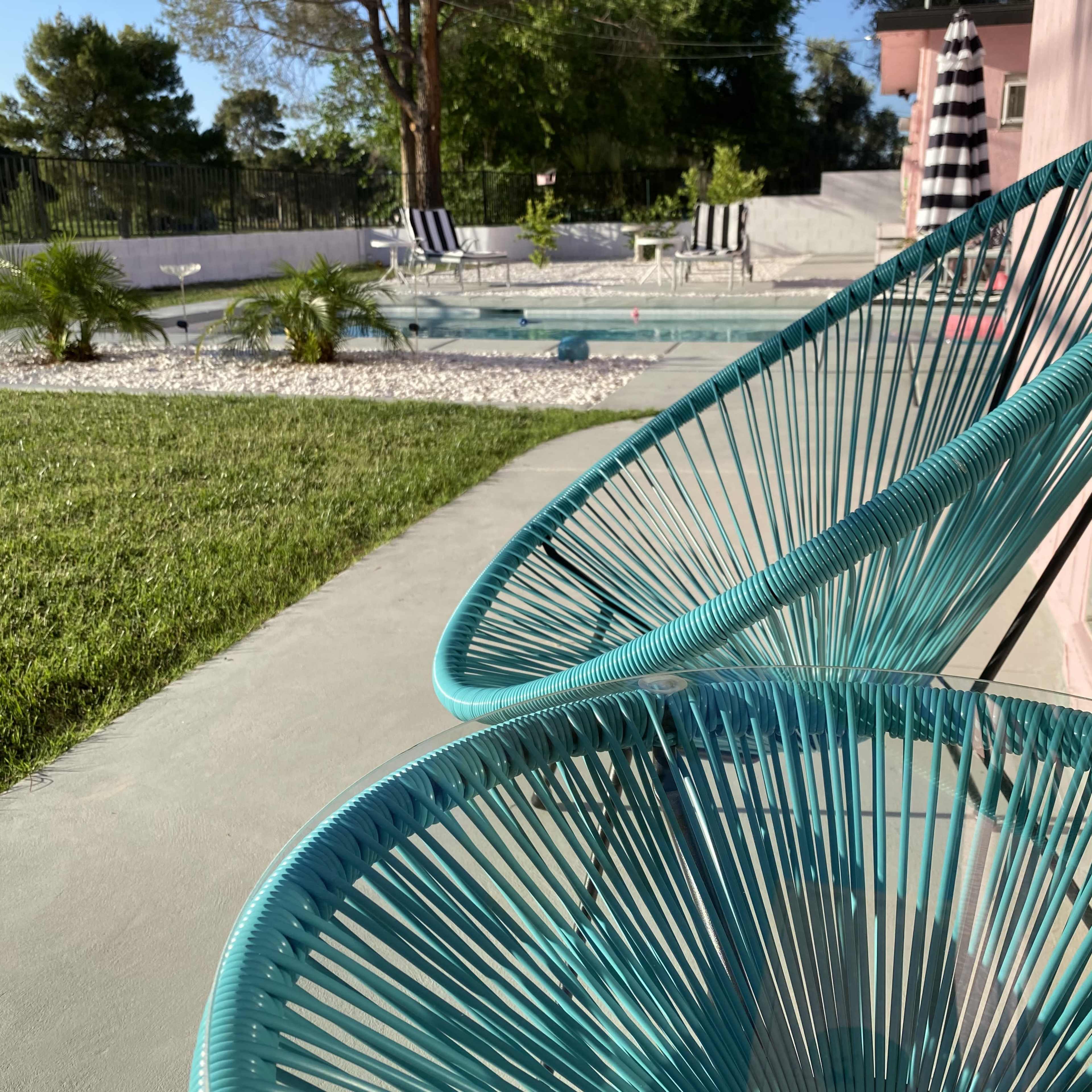 A pair of turquoise chairs are positioned beside a pool area surrounded by greenery and lounge chairs.