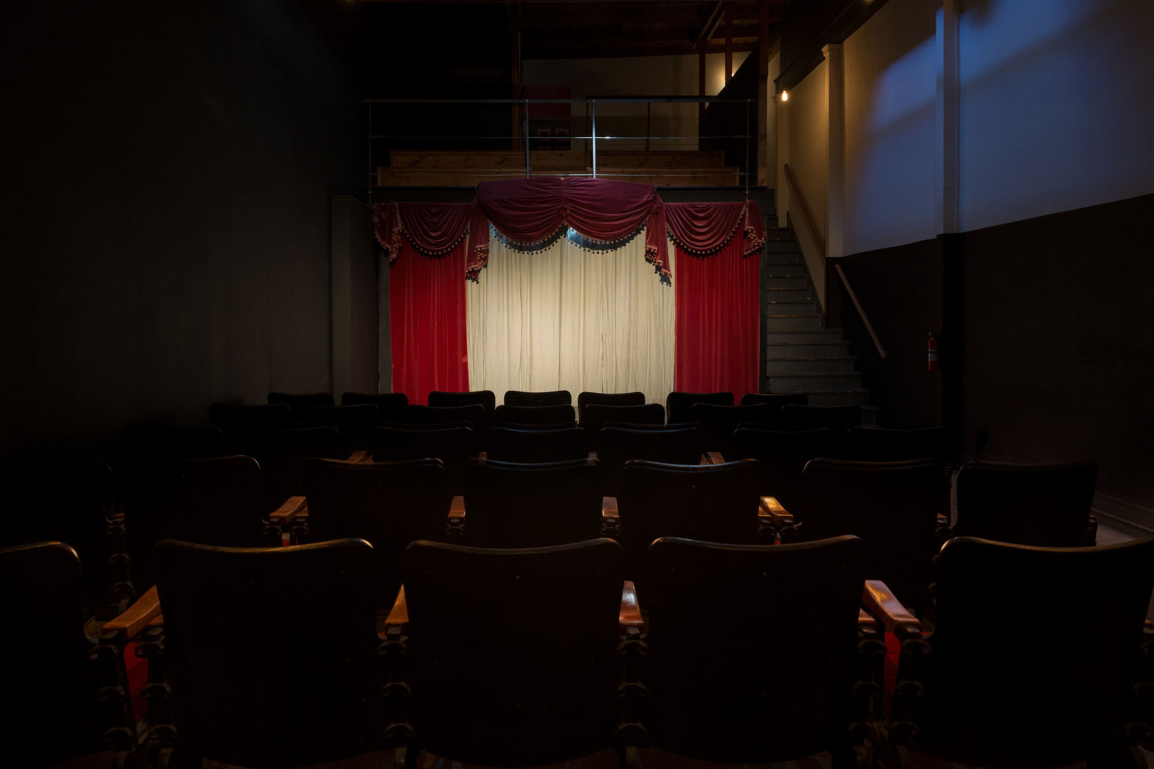 A small theater interior features rows of black audience chairs facing a stage adorned with red curtains and a white backdrop.