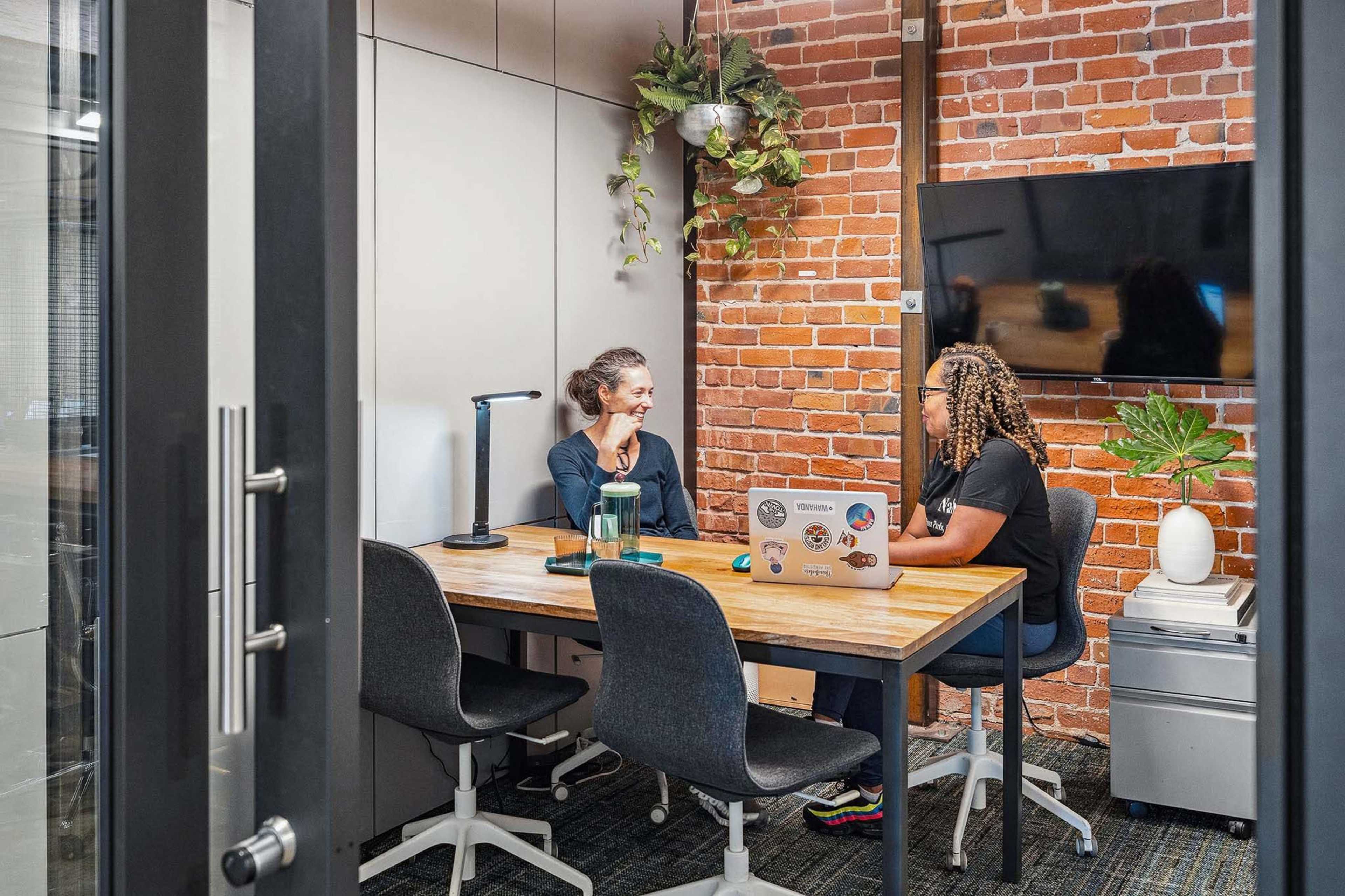 Two women are seated at a wooden table in a small office with exposed brick walls, discussing while using a laptop.