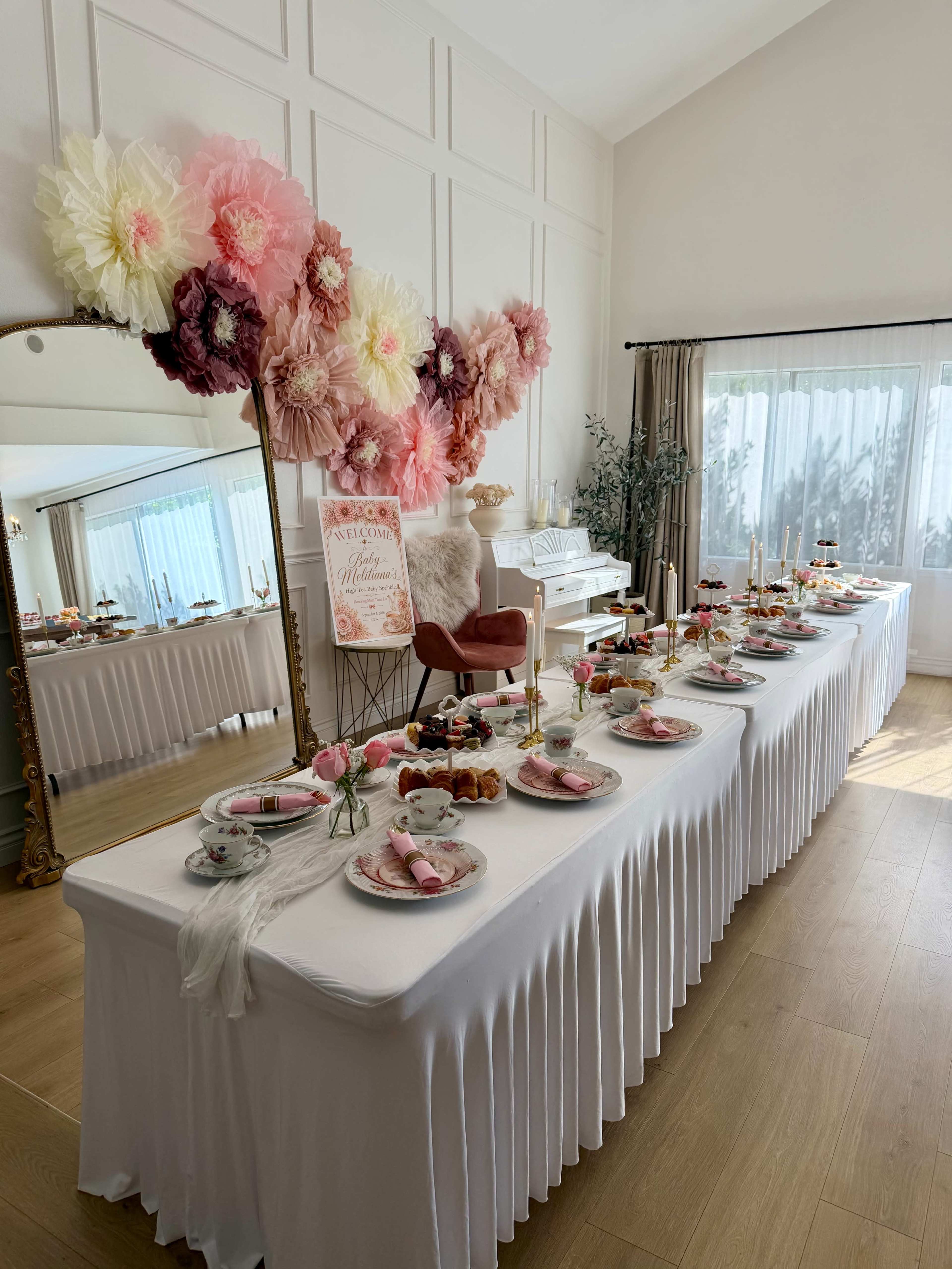 The image shows a beautifully arranged table set for an event, featuring a variety of desserts, floral decorations, and a decorative sign in a well-lit room.
