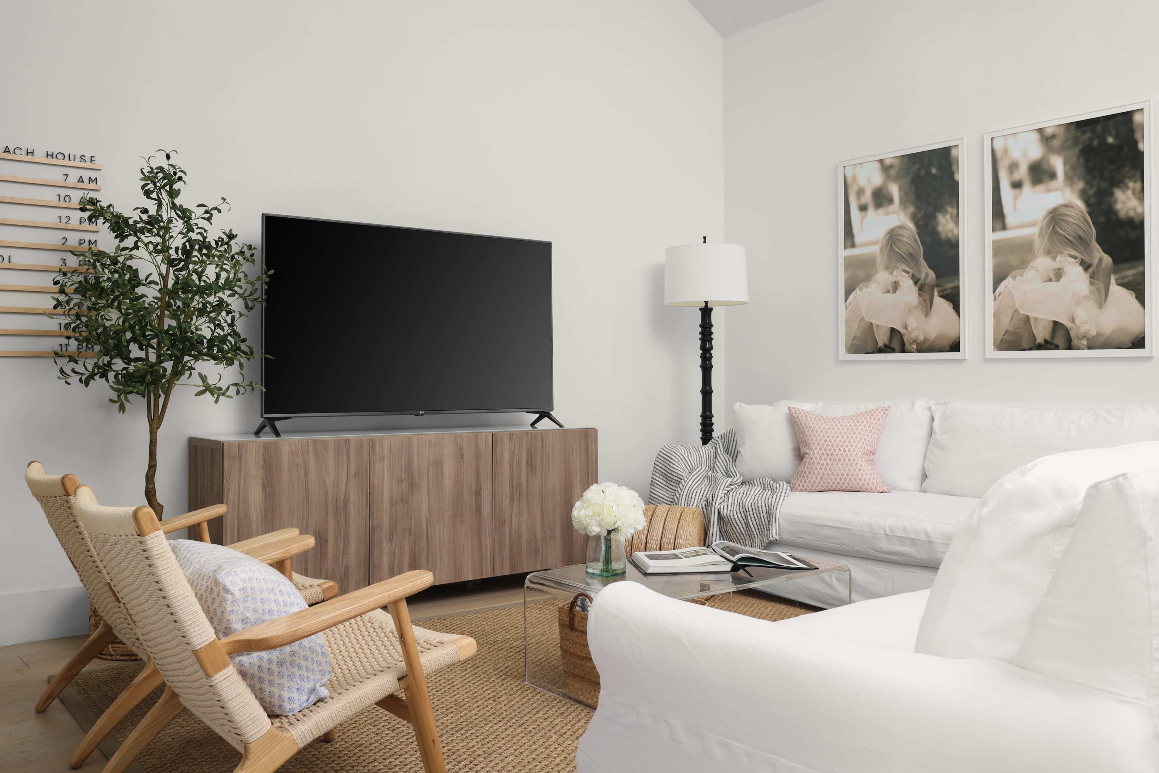 A modern living room features a large TV on a wooden console, surrounded by cozy seating, a lamp, and framed pictures on the wall.