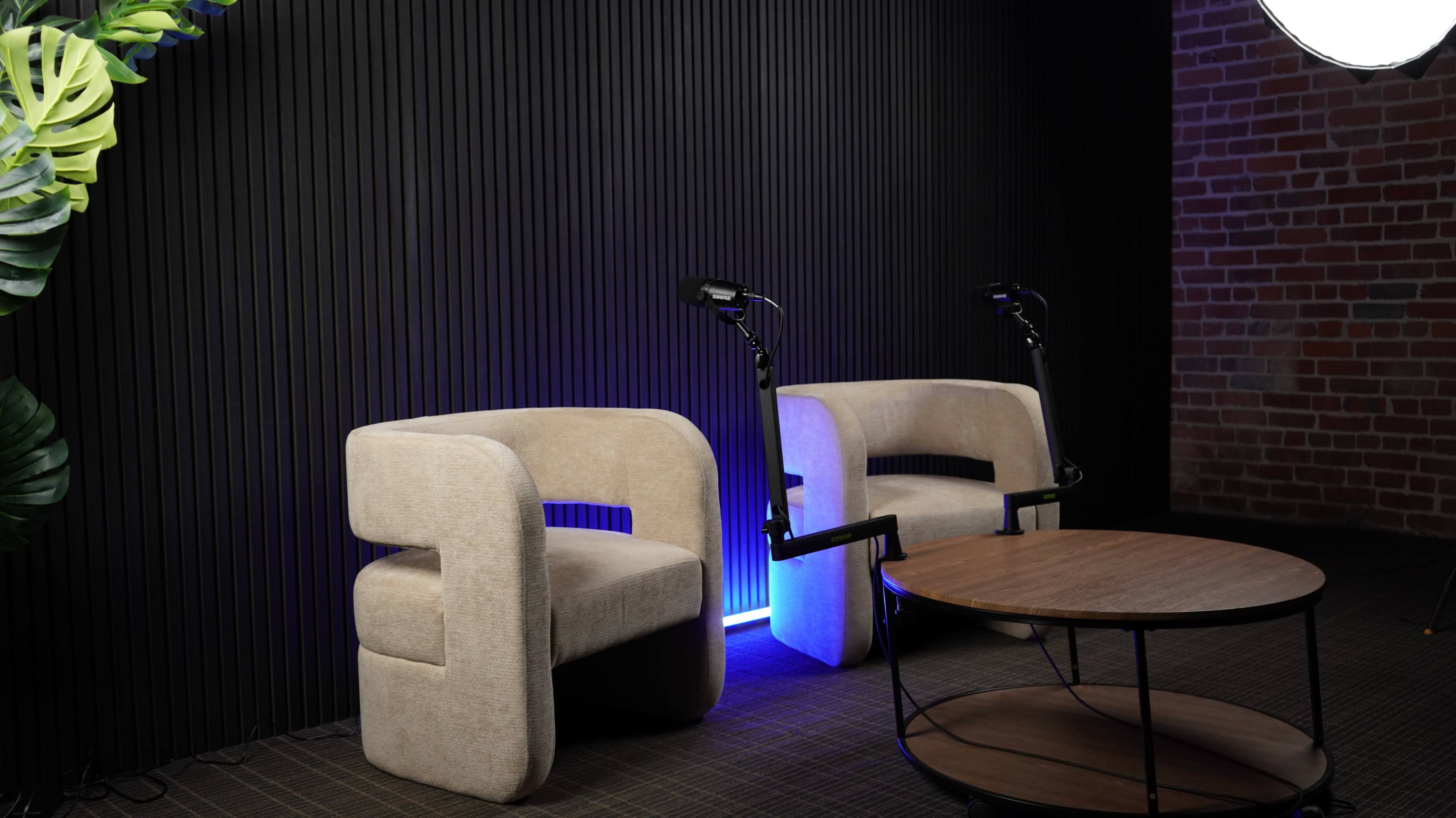 The image shows two beige chairs positioned beside a round wooden table, with a dark textured wall and a light source creating a blue glow behind them.