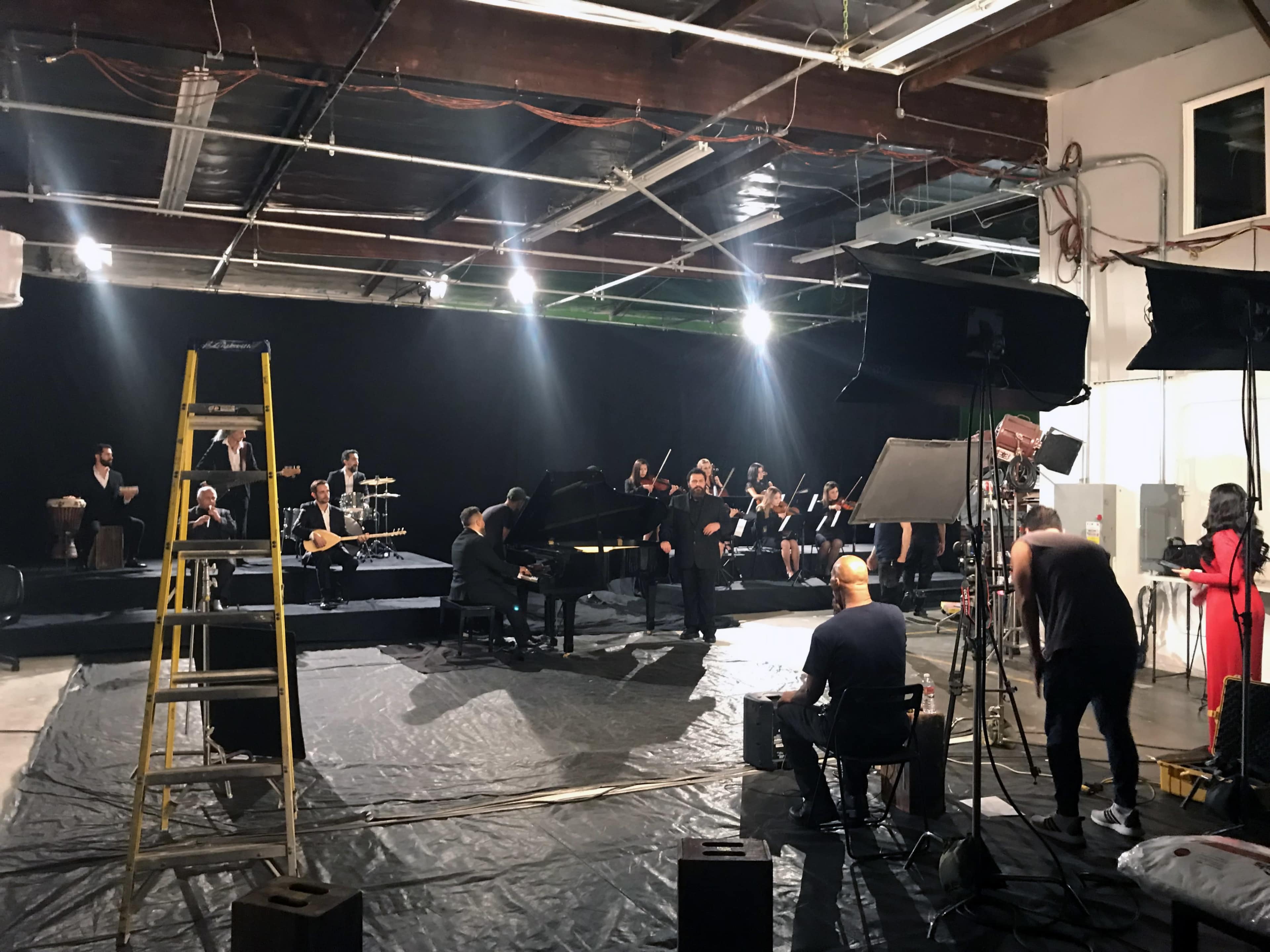 A large group of musicians performs on a soundstage, with a grand piano at the center and various instruments and equipment arranged around them.