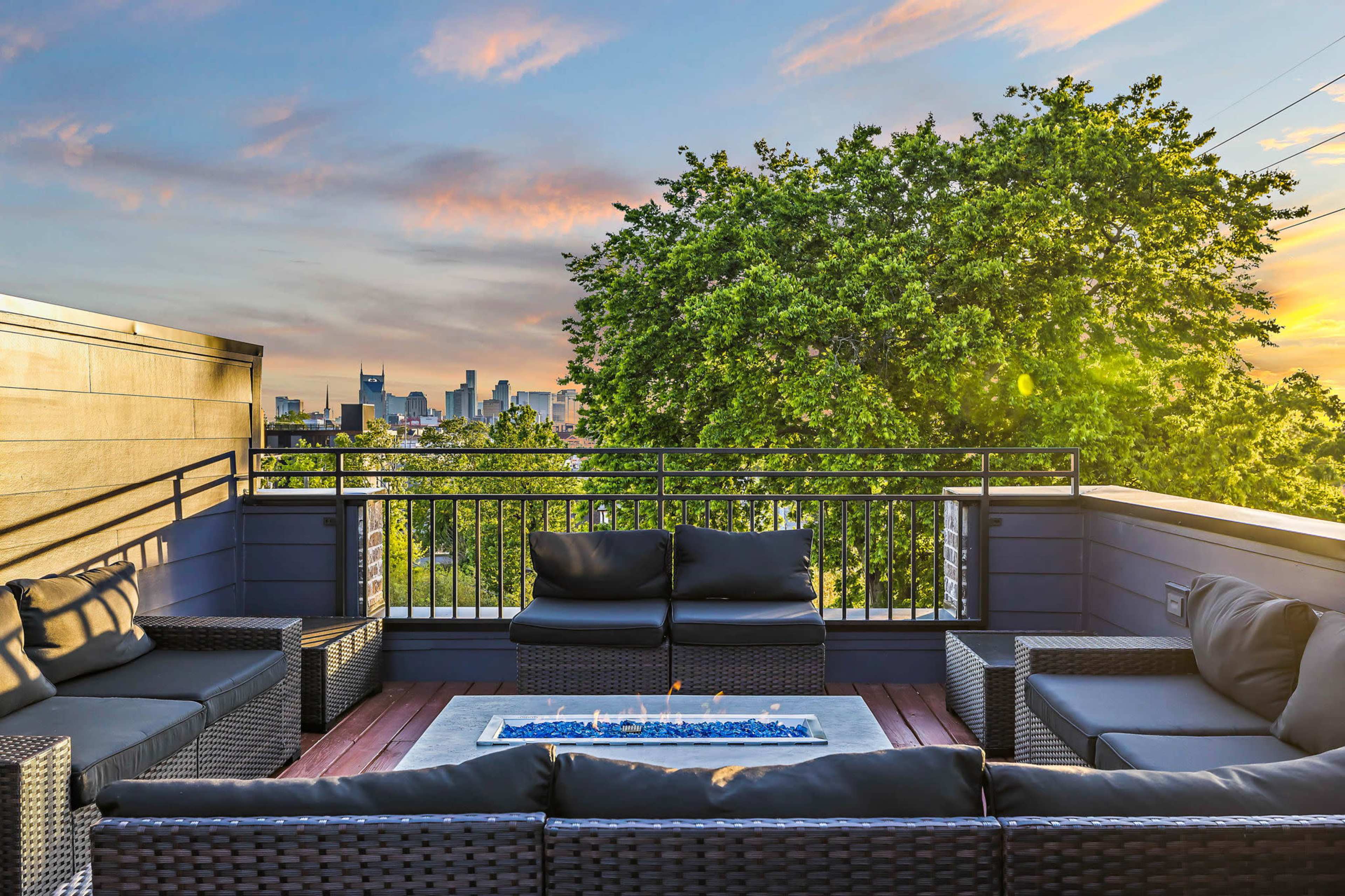 The image shows a rooftop patio with a sectional couch and a fire table, overlooking a city skyline and greenery at sunset.