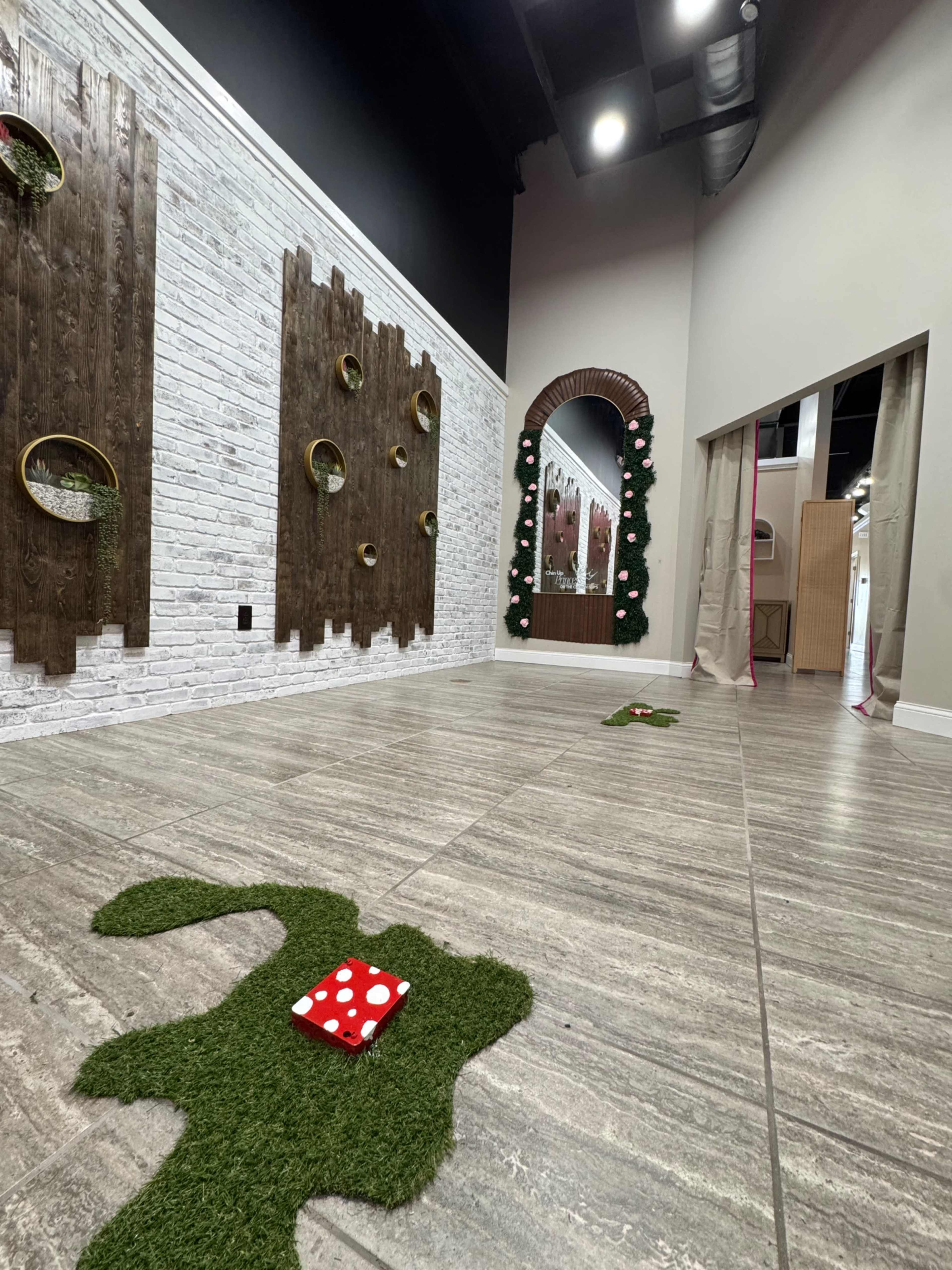 The image shows an interior room with decorative wooden wall features, faux grass patches on the floor, and a mirror framed with plants.