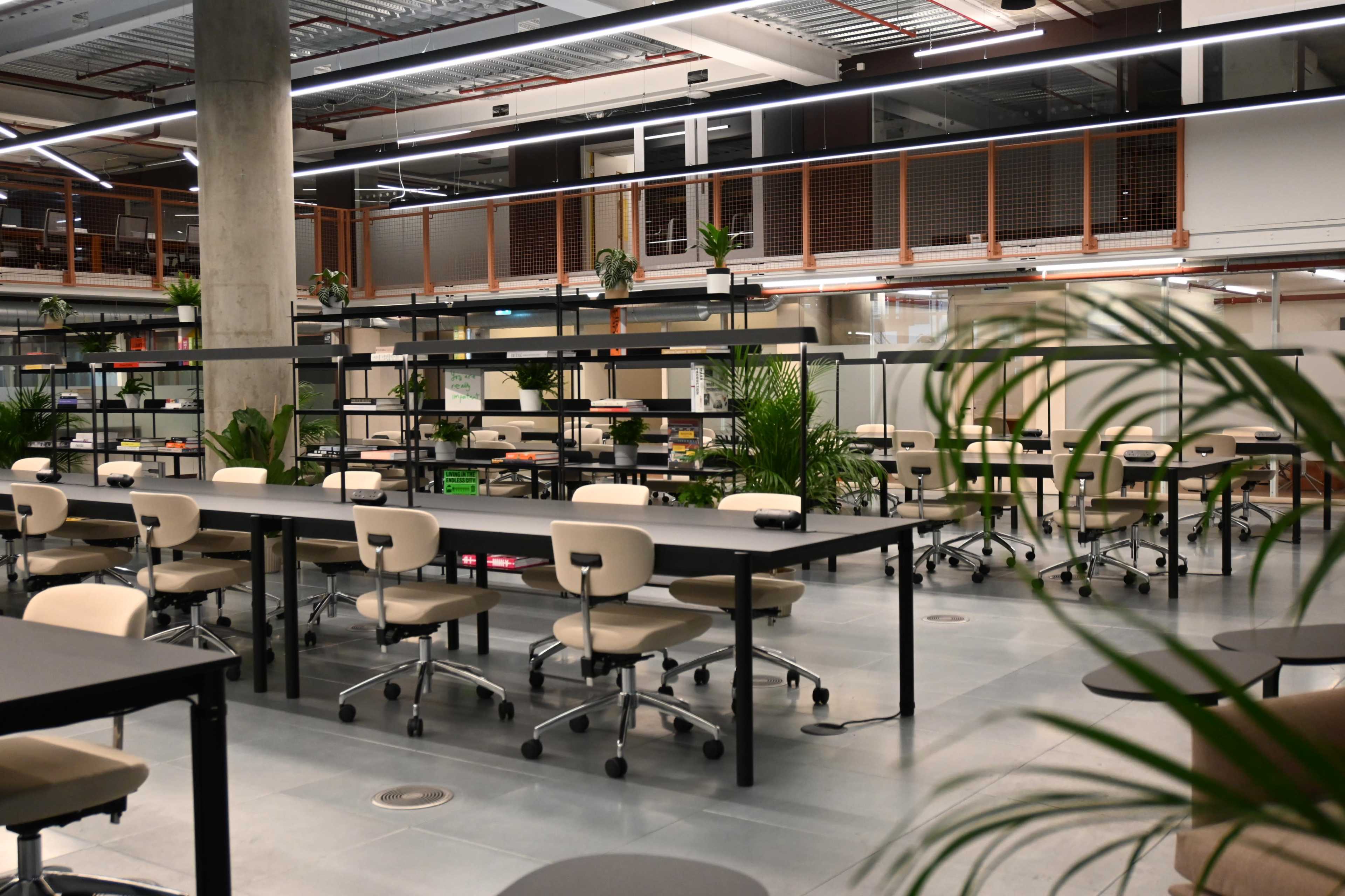 The image shows a modern office space with several tables, rolling chairs, and indoor plants, all arranged in an open layout.