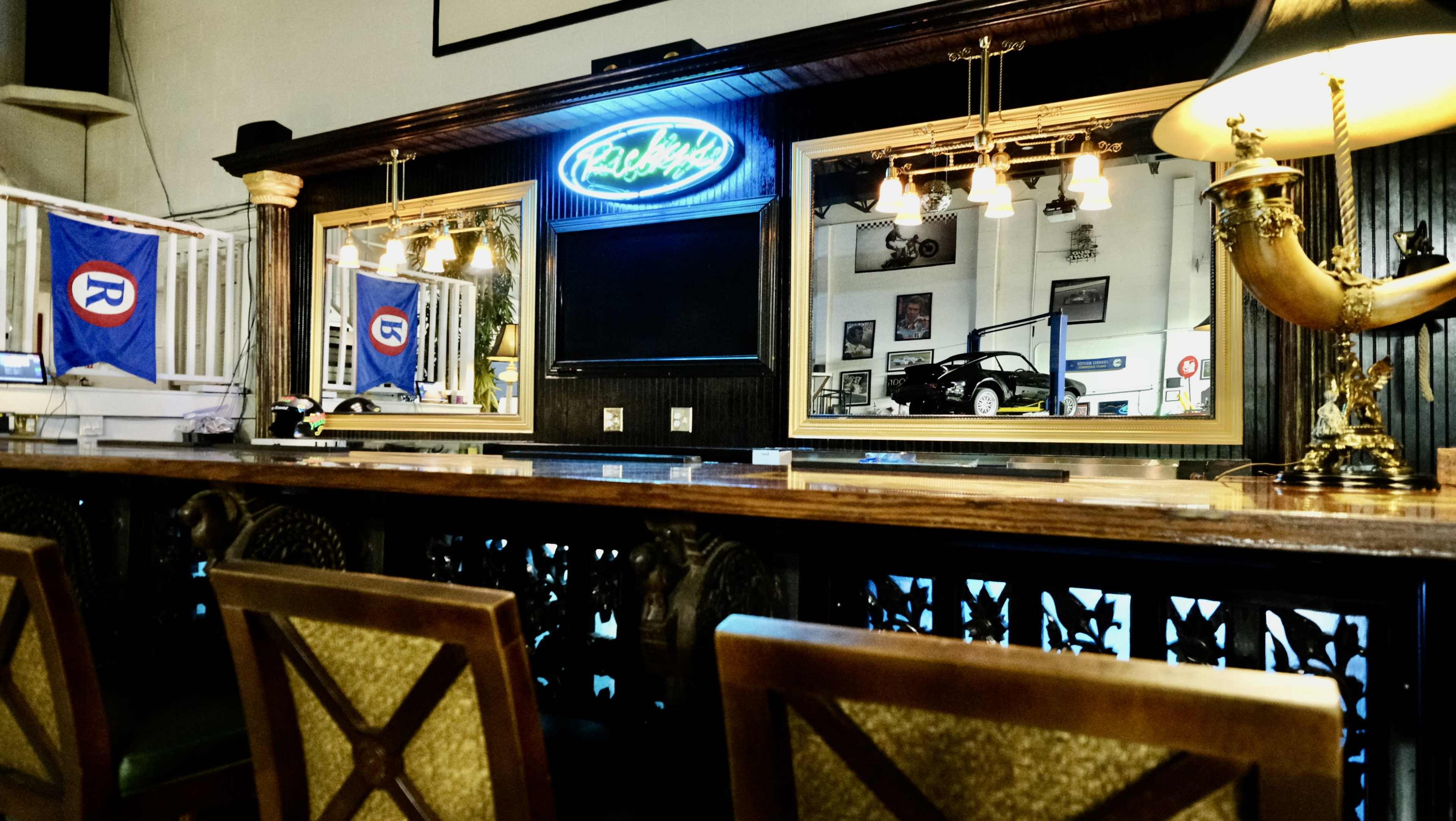 A bar area with a wooden counter, illuminated mirrors, and a blue neon sign, flanked by framed photographs and a few flags hanging nearby.