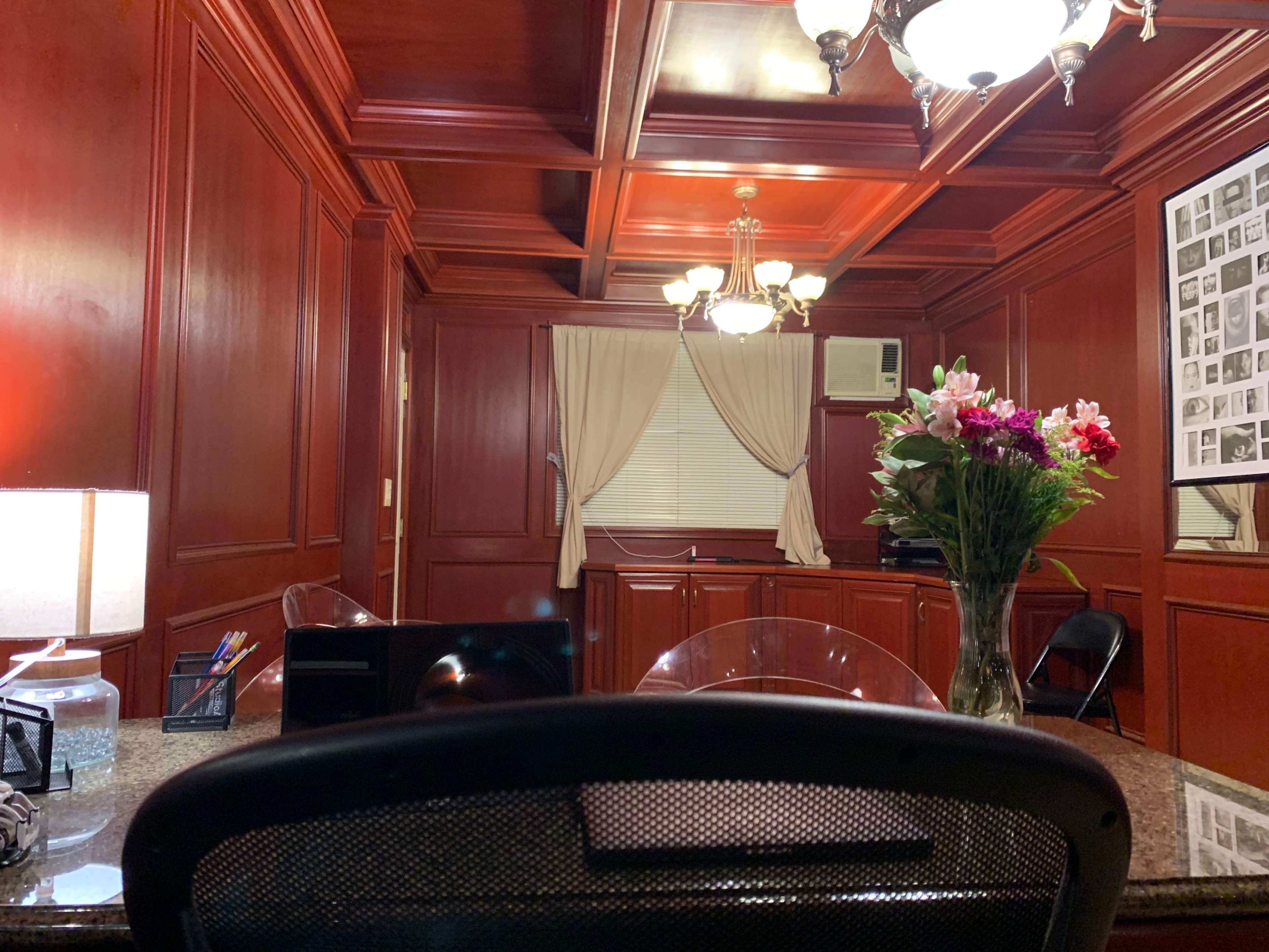 An office with dark wooden paneling, a large desk, a bouquet of flowers, and a window covered by a curtain.
