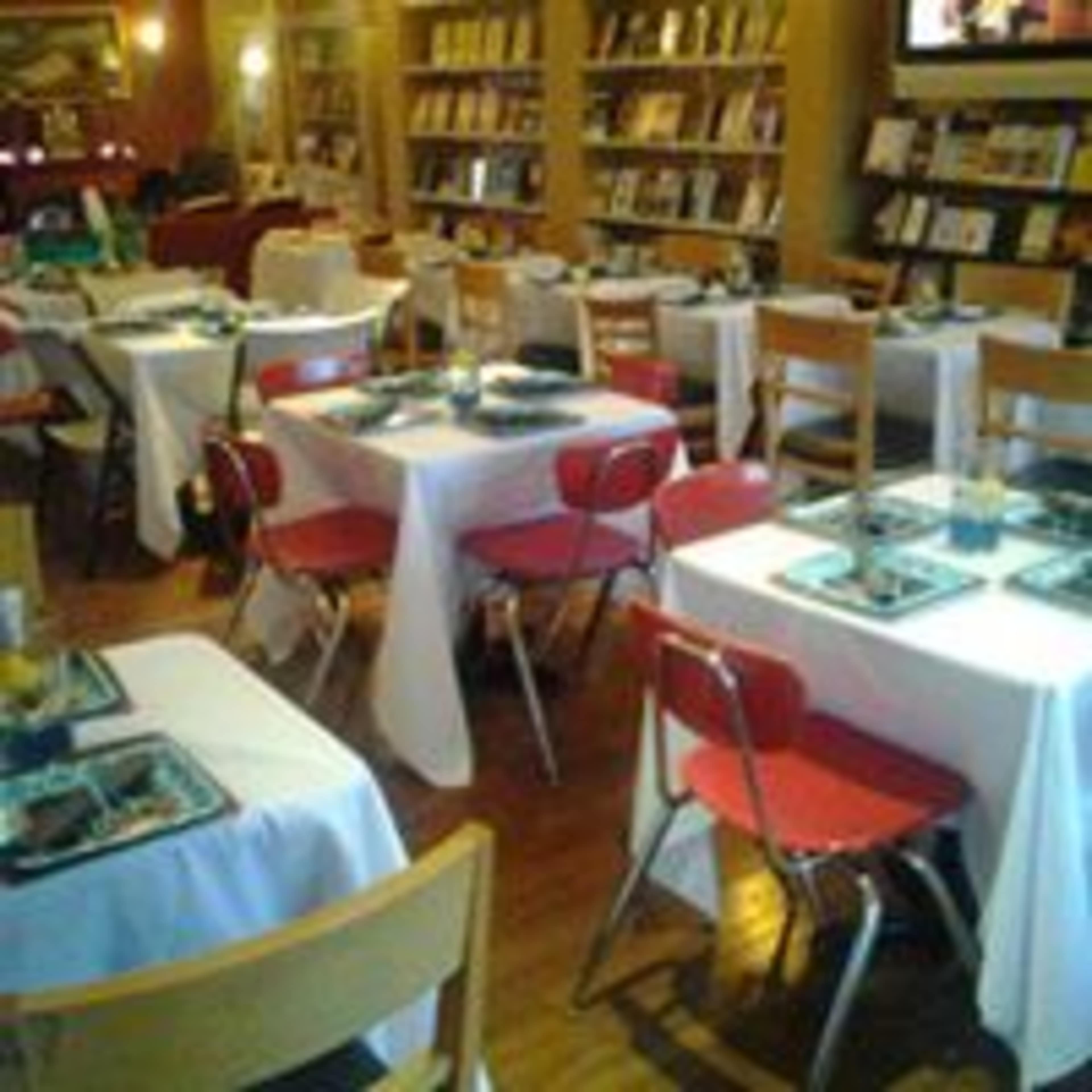 A restaurant interior with white tablecloths covering several tables, each set with plates and drinks, surrounded by shelves filled with books.