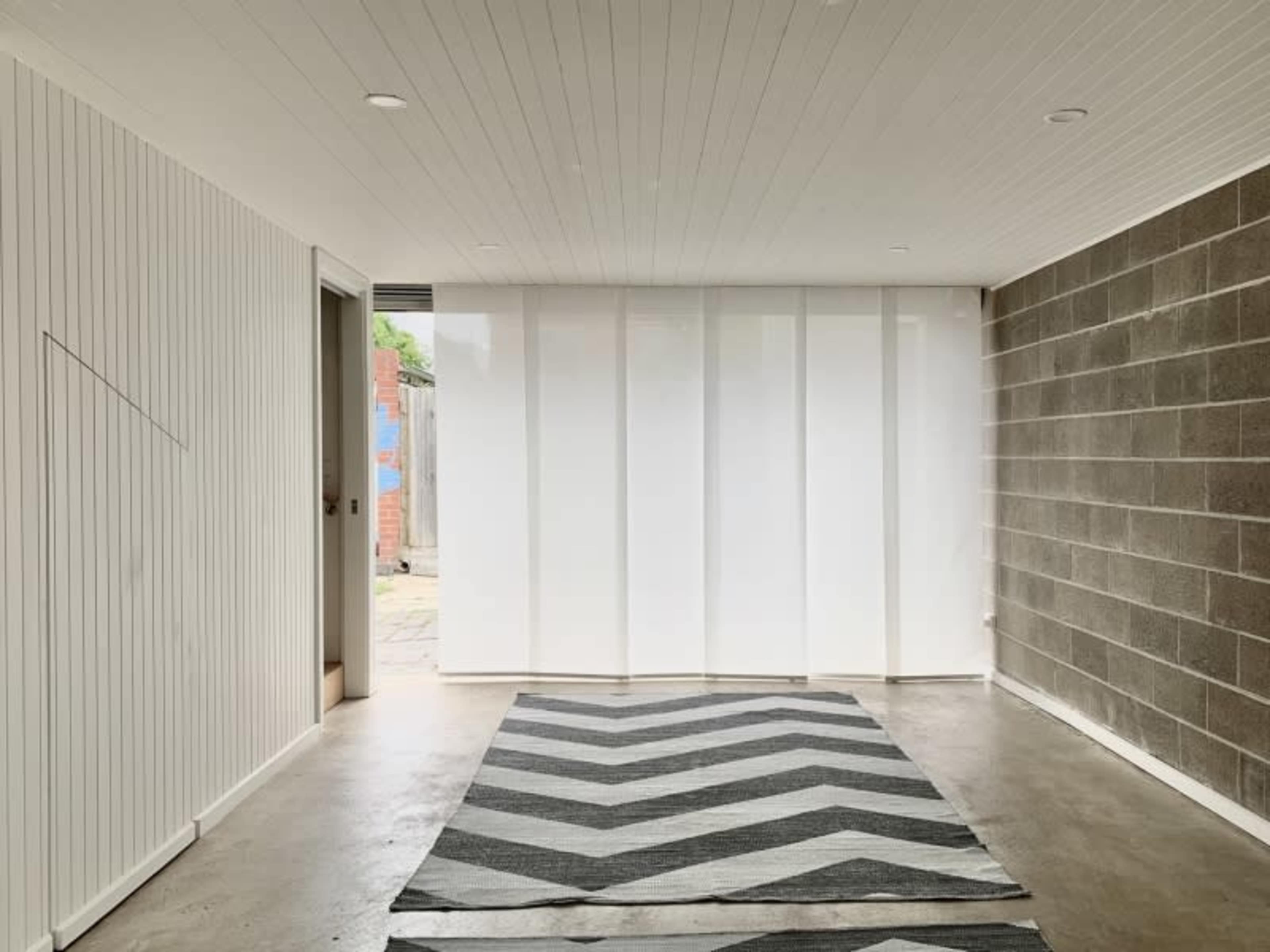 A minimalist interior space with a concrete wall and a white, striped fabric partition, featuring a chevron-patterned rug on the floor.