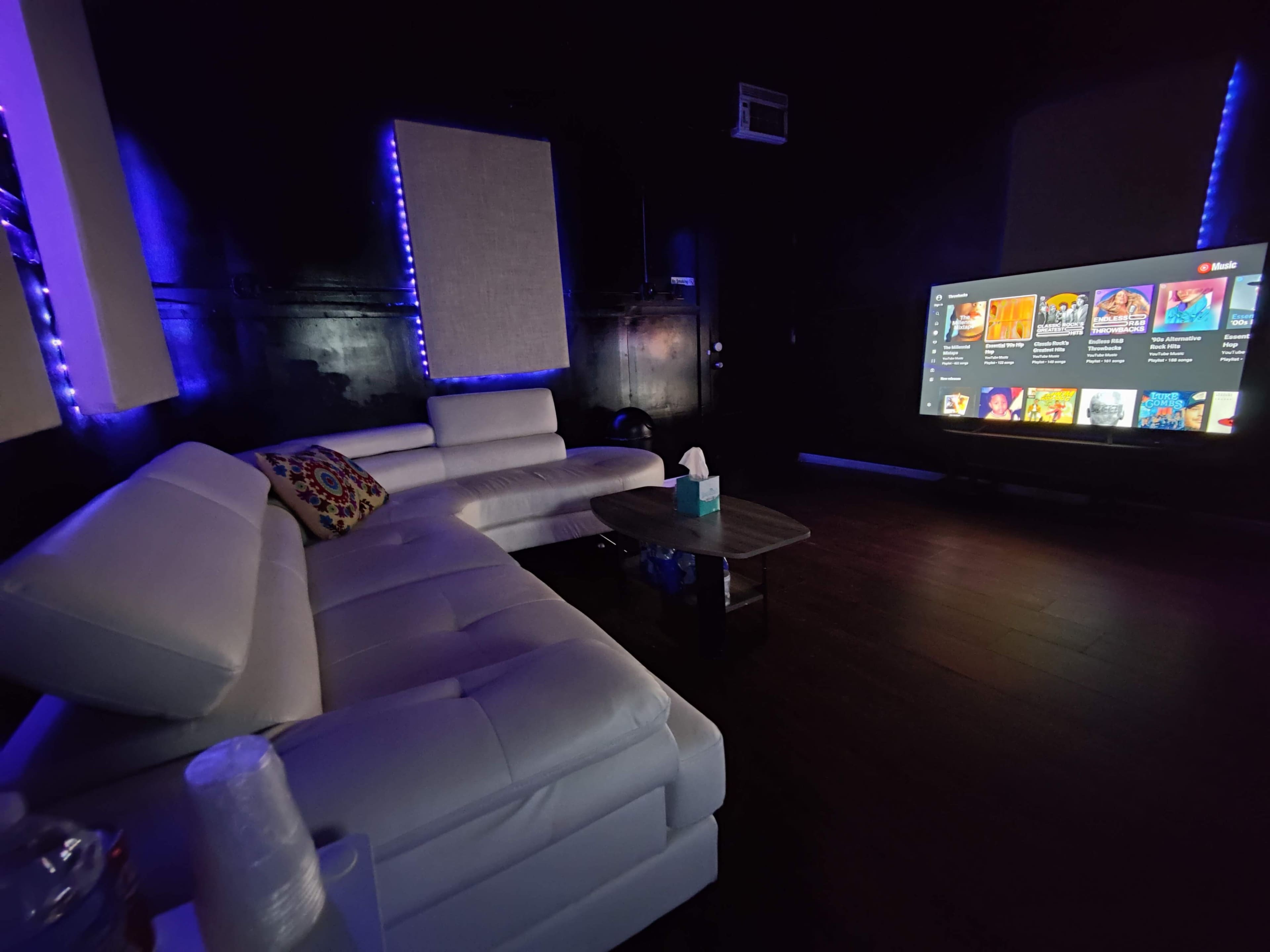 The image shows a dimly lit room featuring a modern white sectional sofa, a small table with a tissue box, and a large television displaying a menu interface.