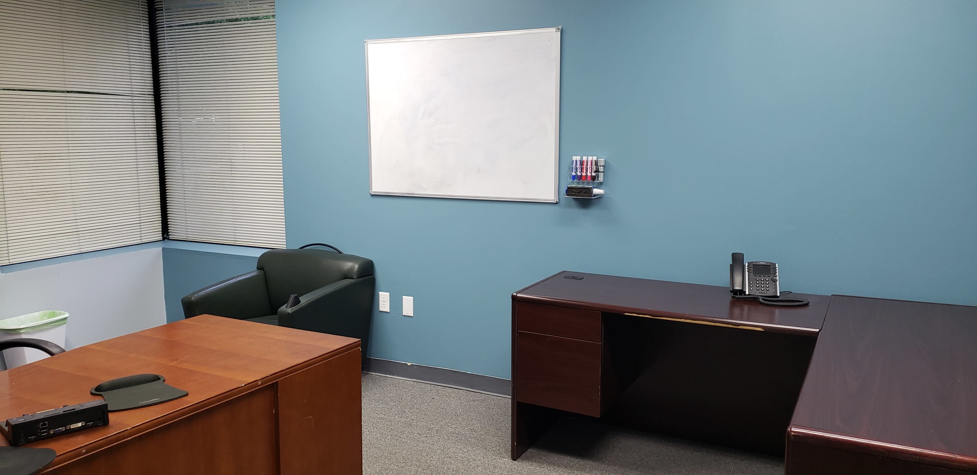 A small office with two desks, a green chair, a whiteboard on a blue wall, and a telephone on one desk.