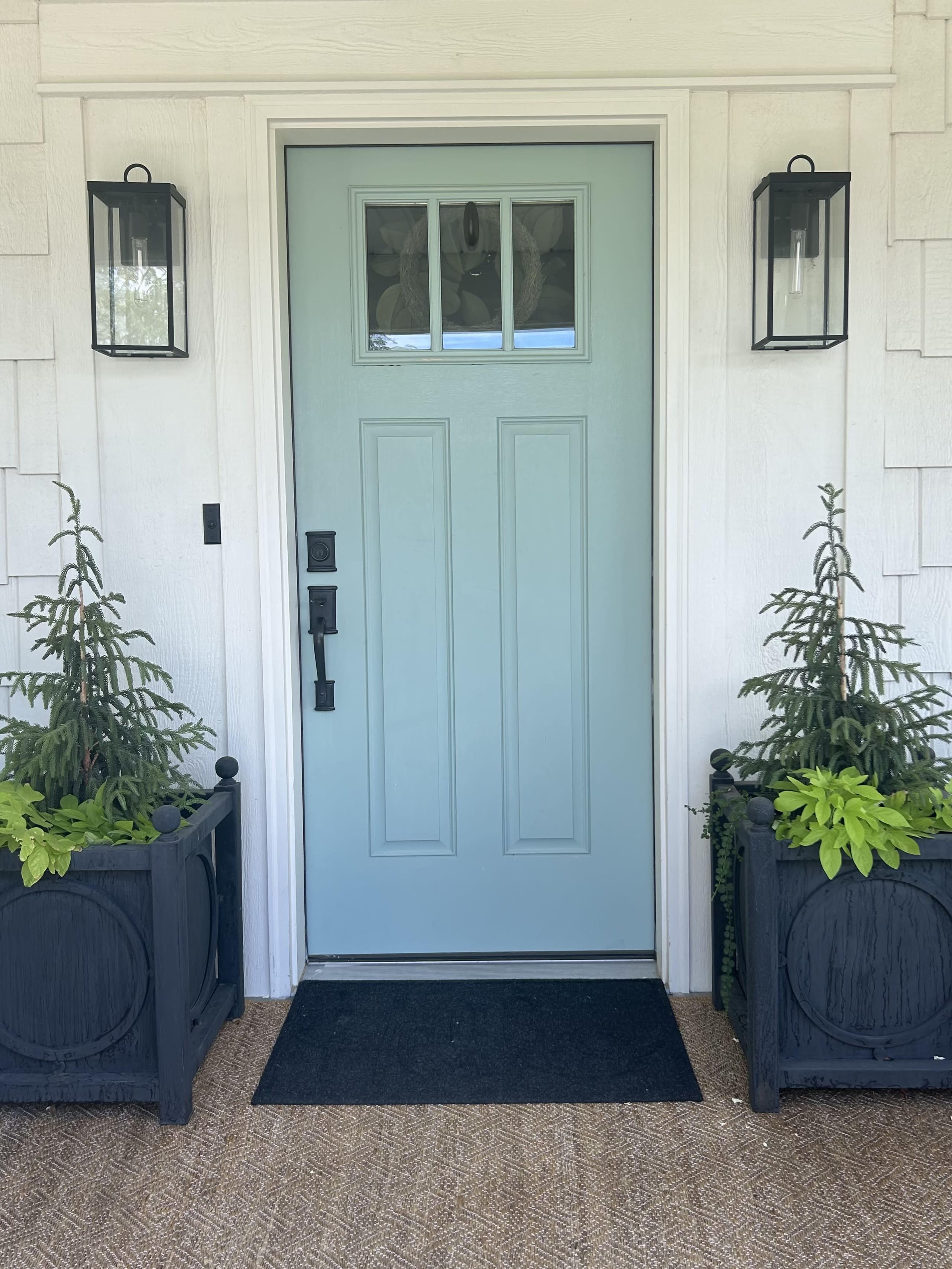 A light blue front door is centered in a white-paneled entryway, flanked by two lantern-style wall sconces and adorned with potted plants on either side.