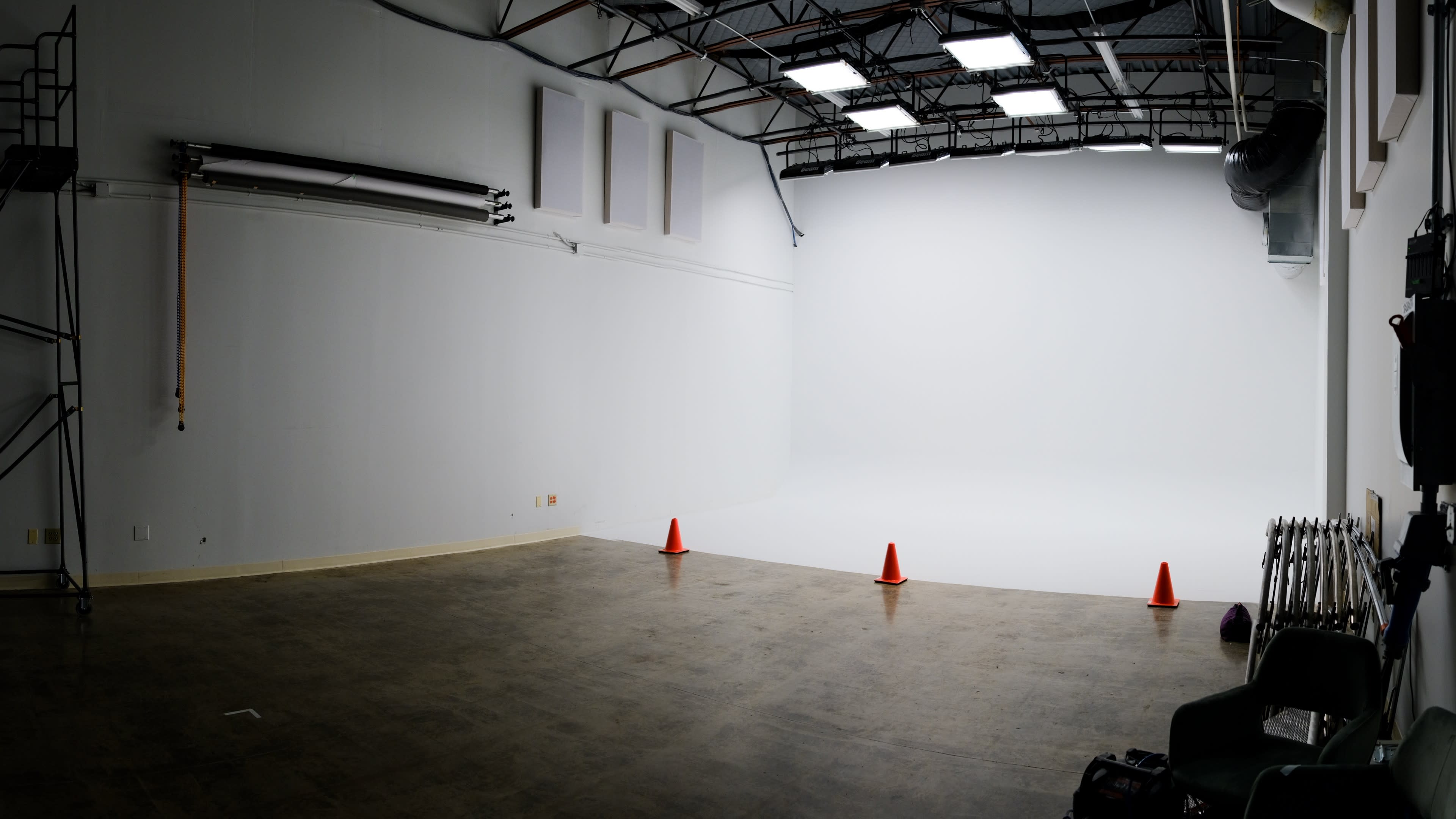 A spacious, empty studio with white walls and a seamless backdrop, illuminated by overhead lights, and featuring three orange traffic cones on the floor.
