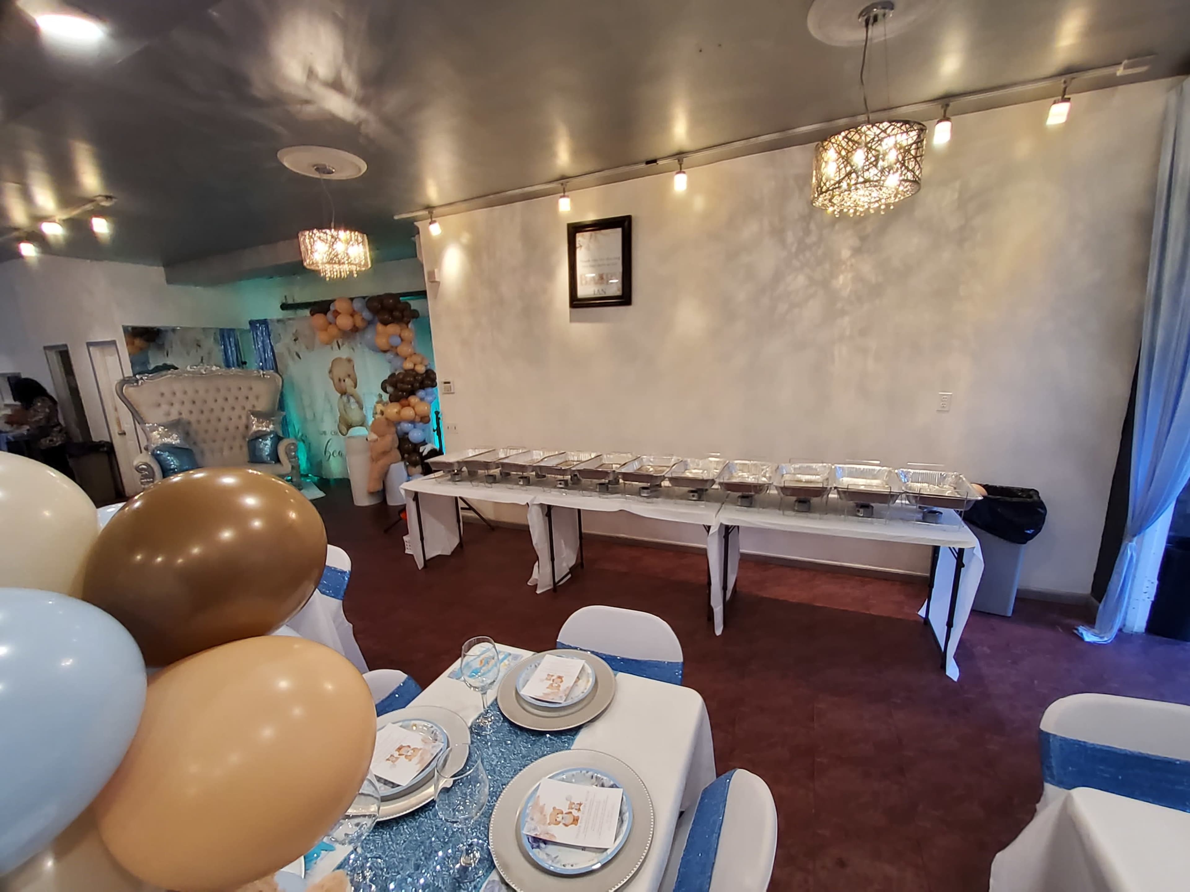 A decorated event space with tables set for a gathering, featuring a buffet line of food and balloons in the foreground.