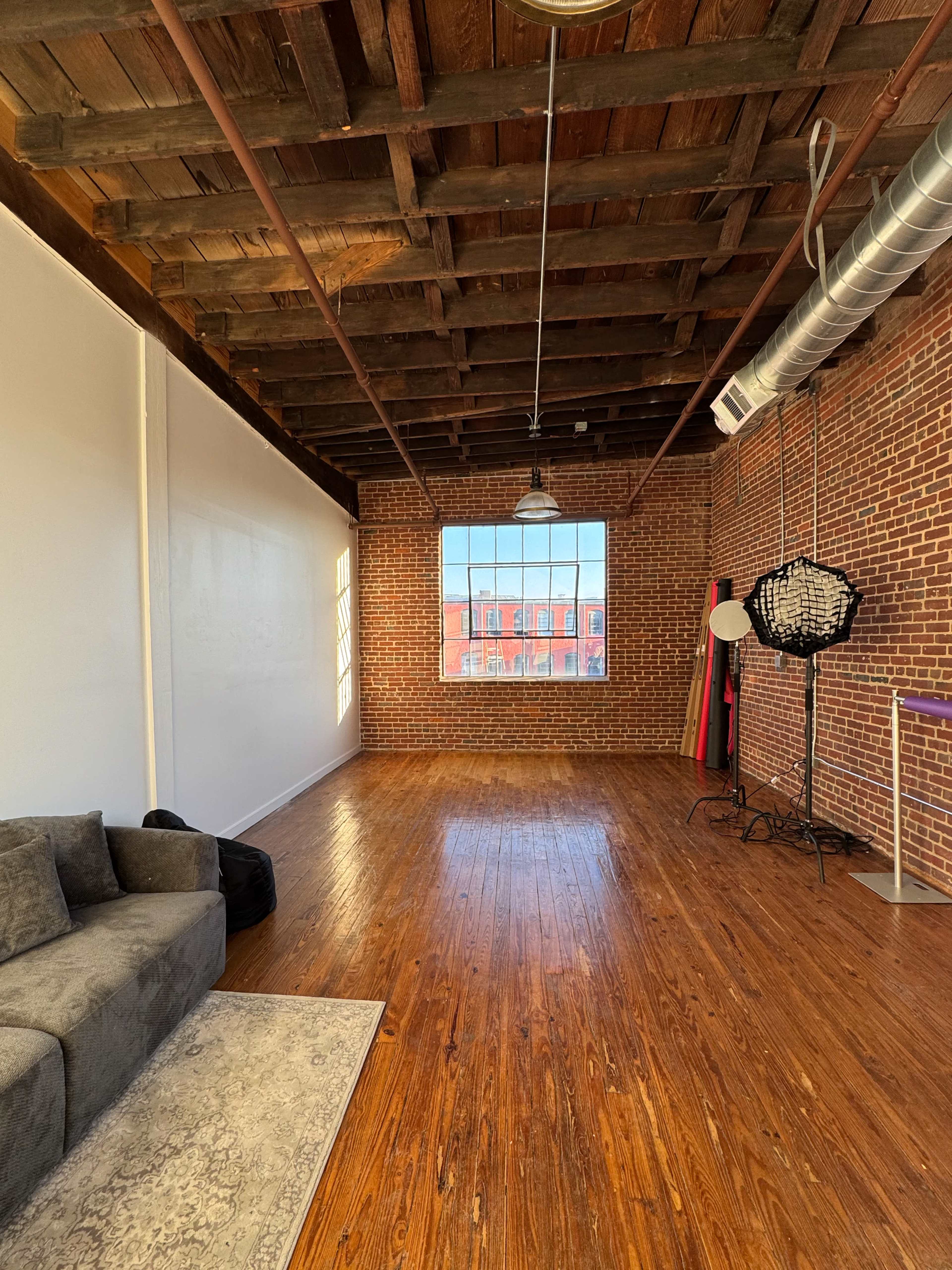 The image shows a spacious room with exposed brick walls, wooden beams on the ceiling, and a hardwood floor, featuring a couch and a large window.