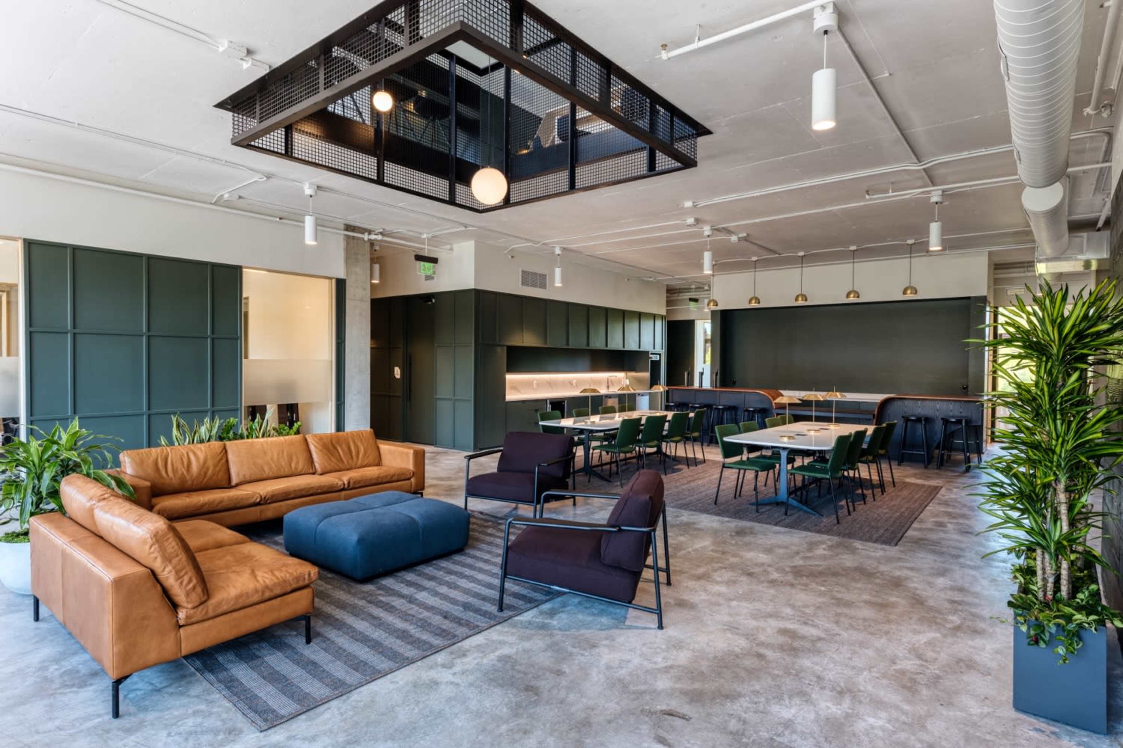 The image shows a modern office space featuring a brown leather sofa, a blue ottoman, a dark dining area, and potted plants.