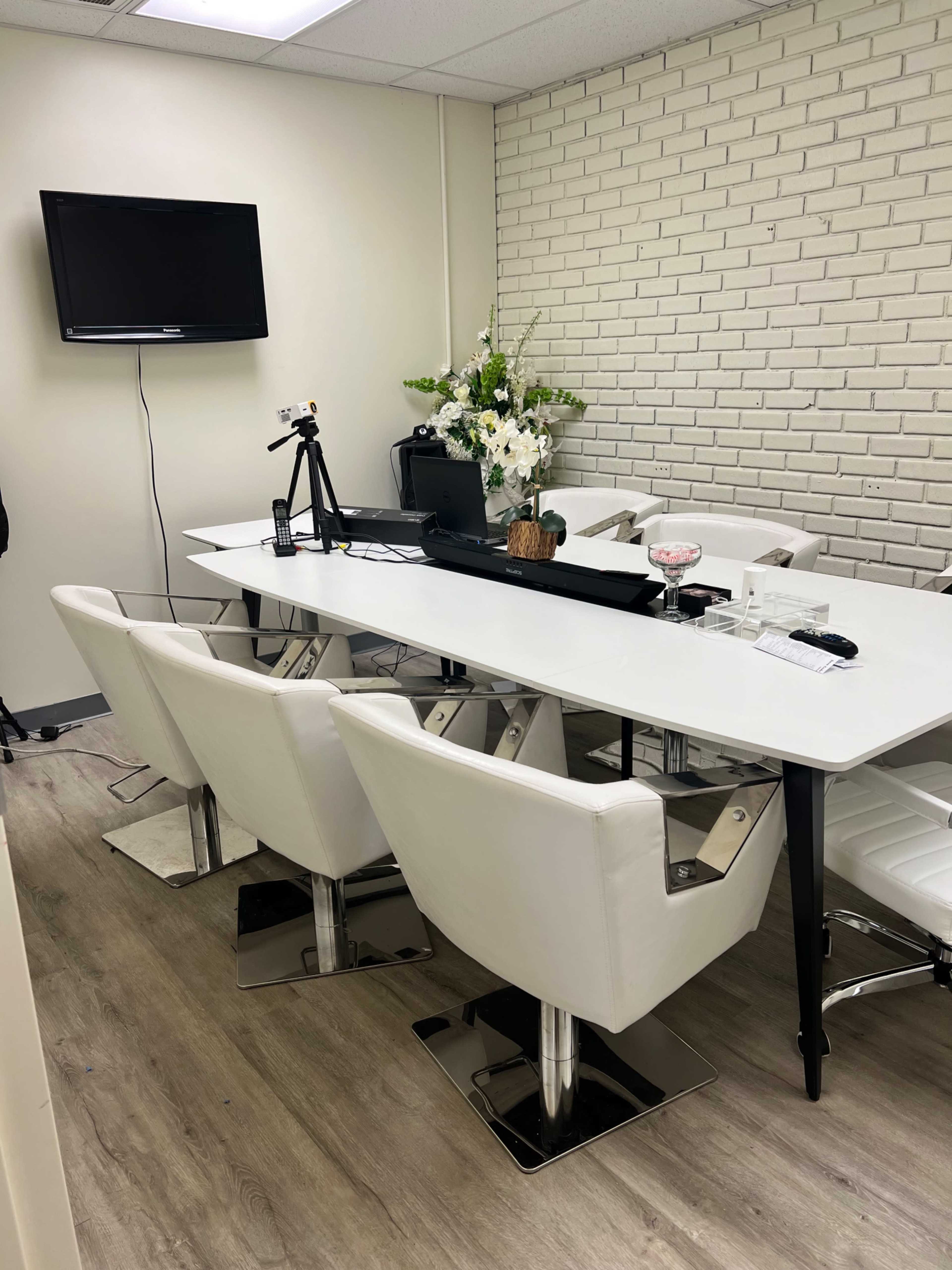 A modern office meeting room features a long white table surrounded by white chairs, with a television on the wall and a floral arrangement on the table.