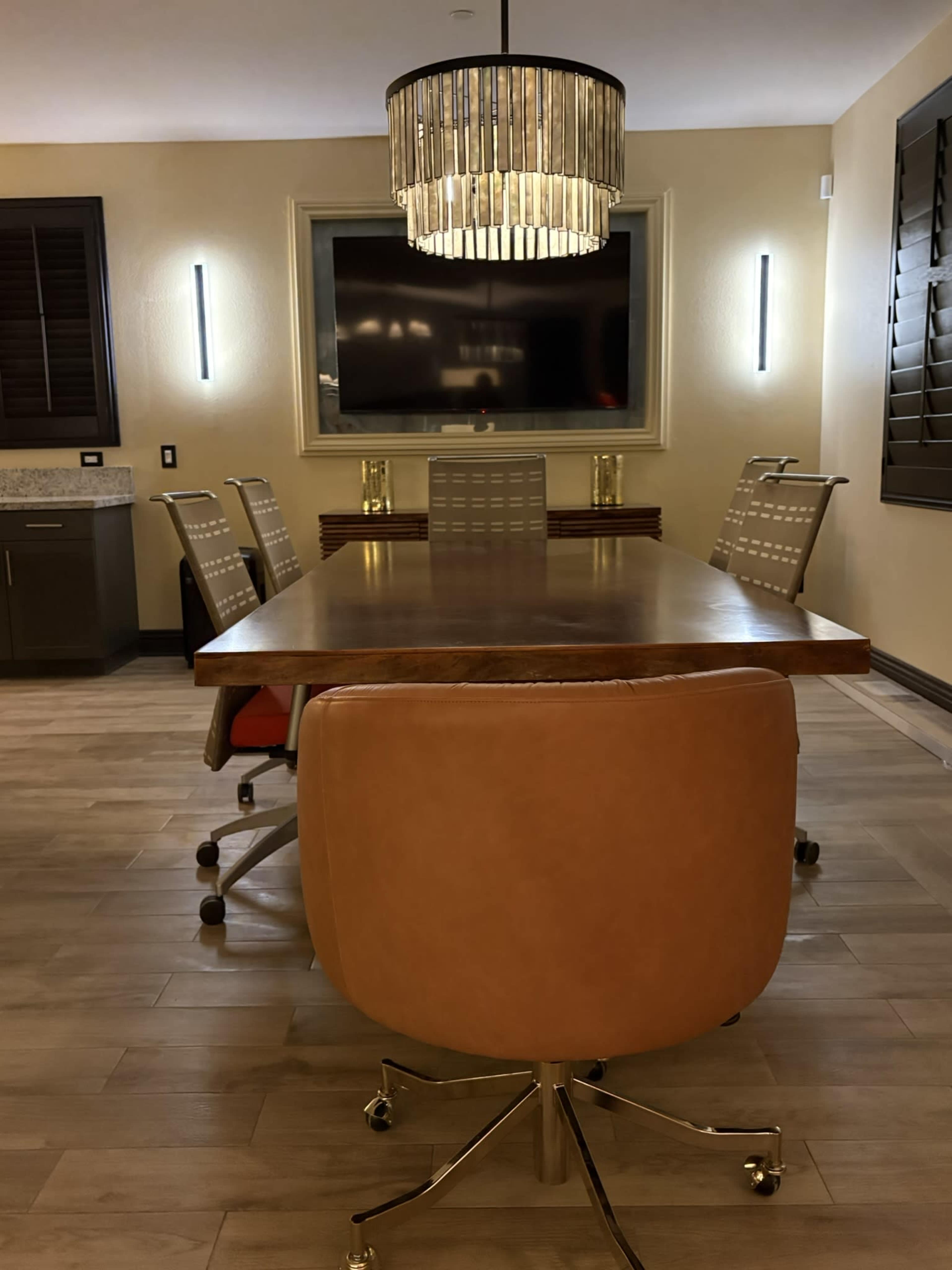 The image shows a modern conference room featuring a long wooden table surrounded by several ergonomic chairs, with a large screen mounted on the wall and a stylish chandelier overhead.