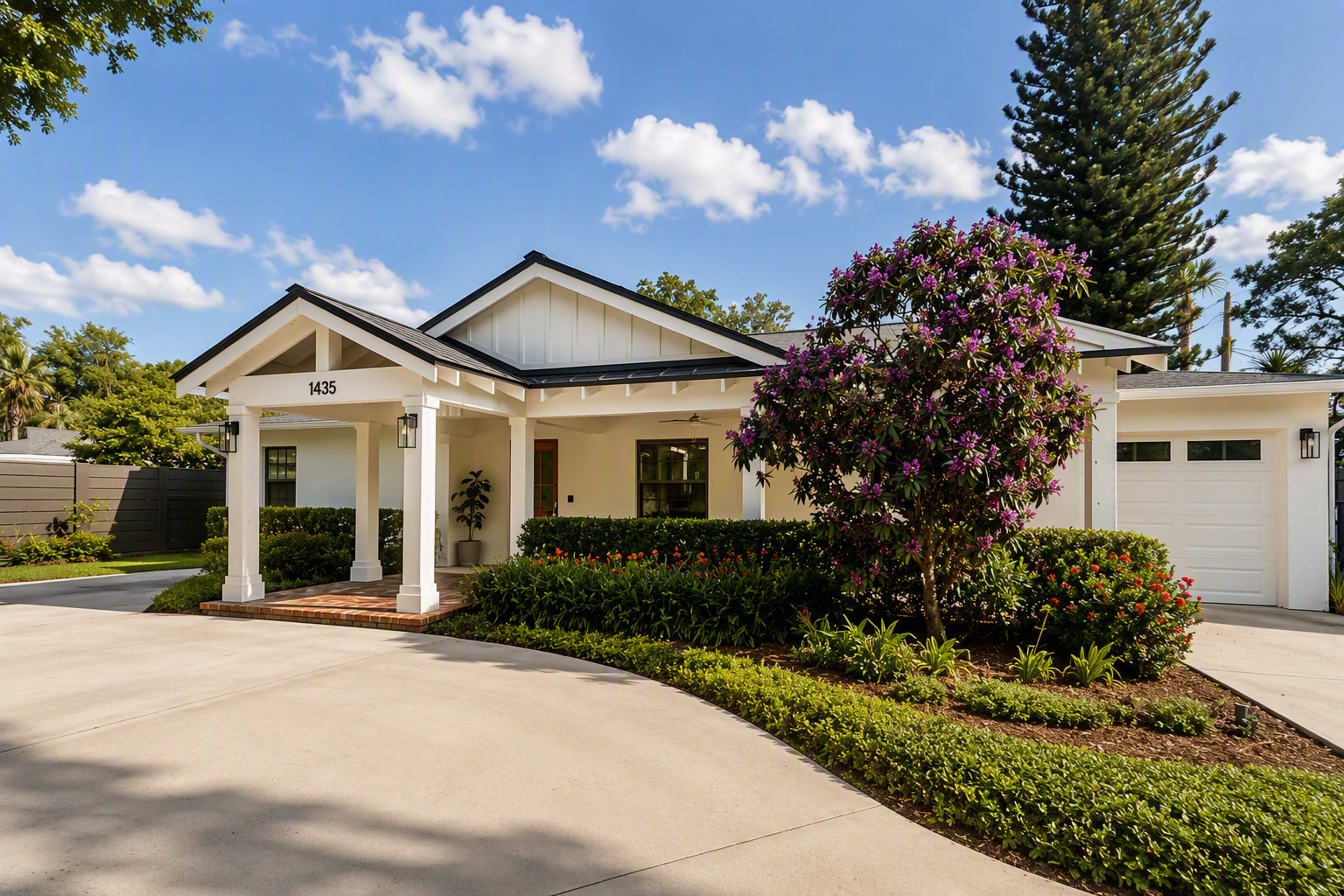 A single-story house with a white exterior and a porch is surrounded by lush green landscaping and a curved driveway.
