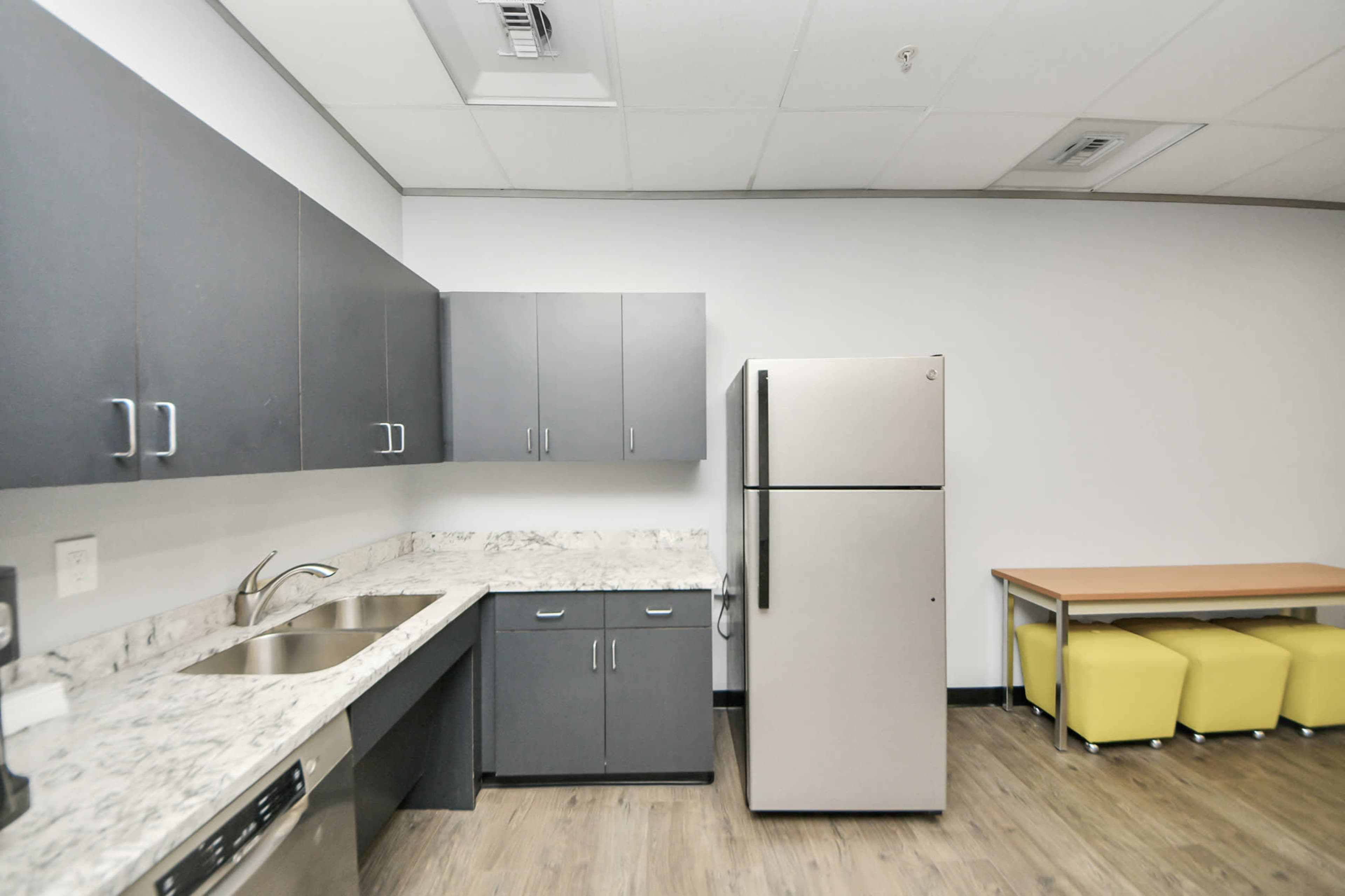 The image shows a modern kitchen with gray cabinets, a stainless steel refrigerator, a sink, and a small dining area with a table and yellow benches.