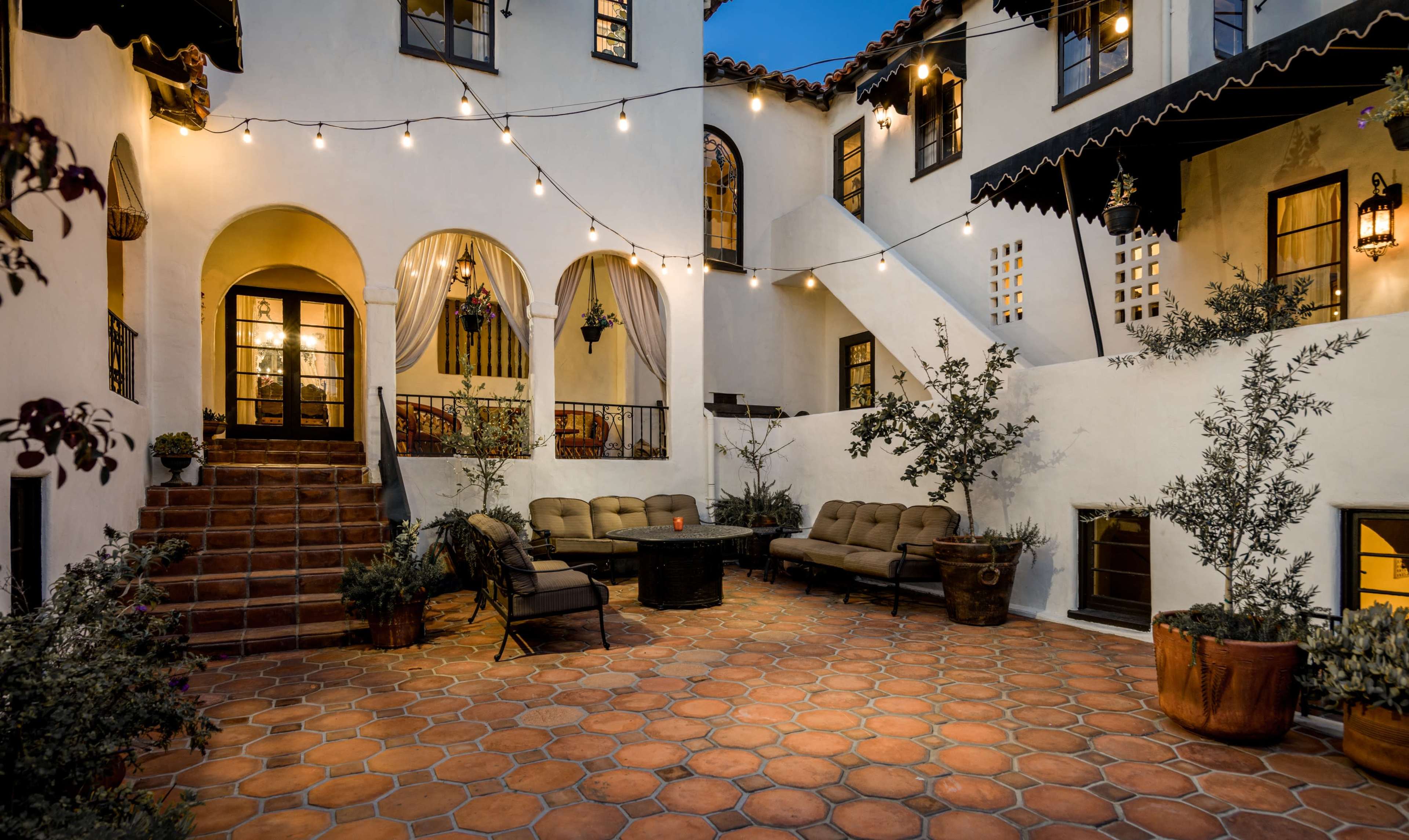 The image depicts a courtyard with terracotta tiles, cozy seating arrangements, potted plants, and string lights illuminating the space.