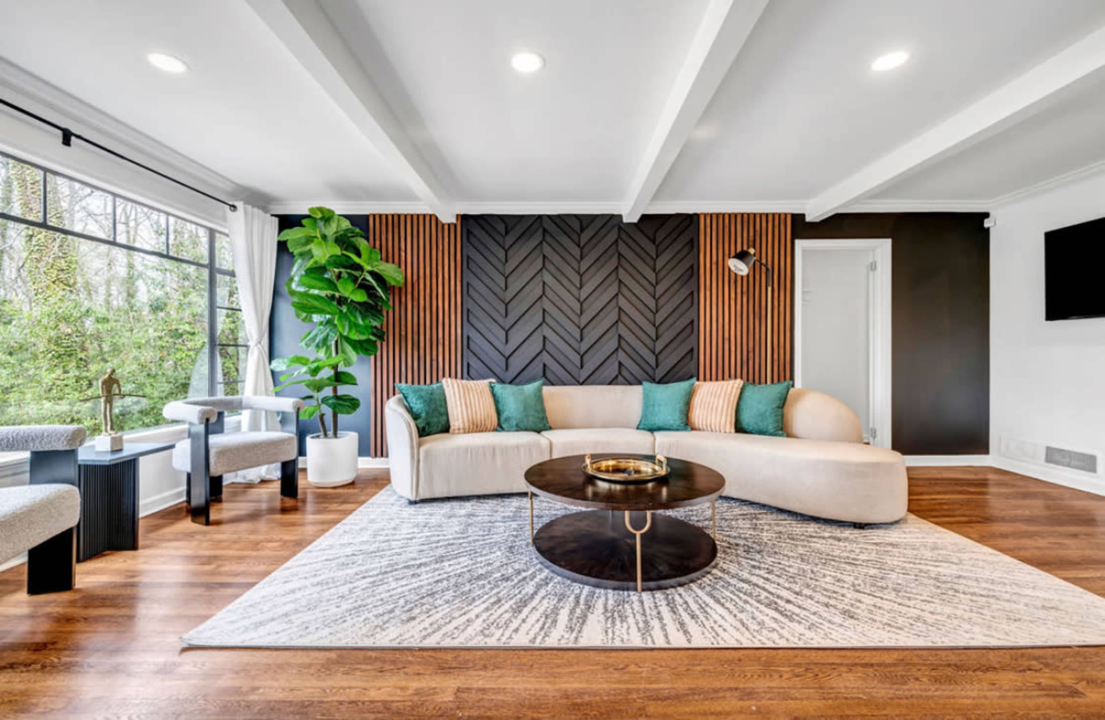 A modern living room features a curved beige couch, dark wooden accents, a round coffee table, and a large window with greenery visible outside.
