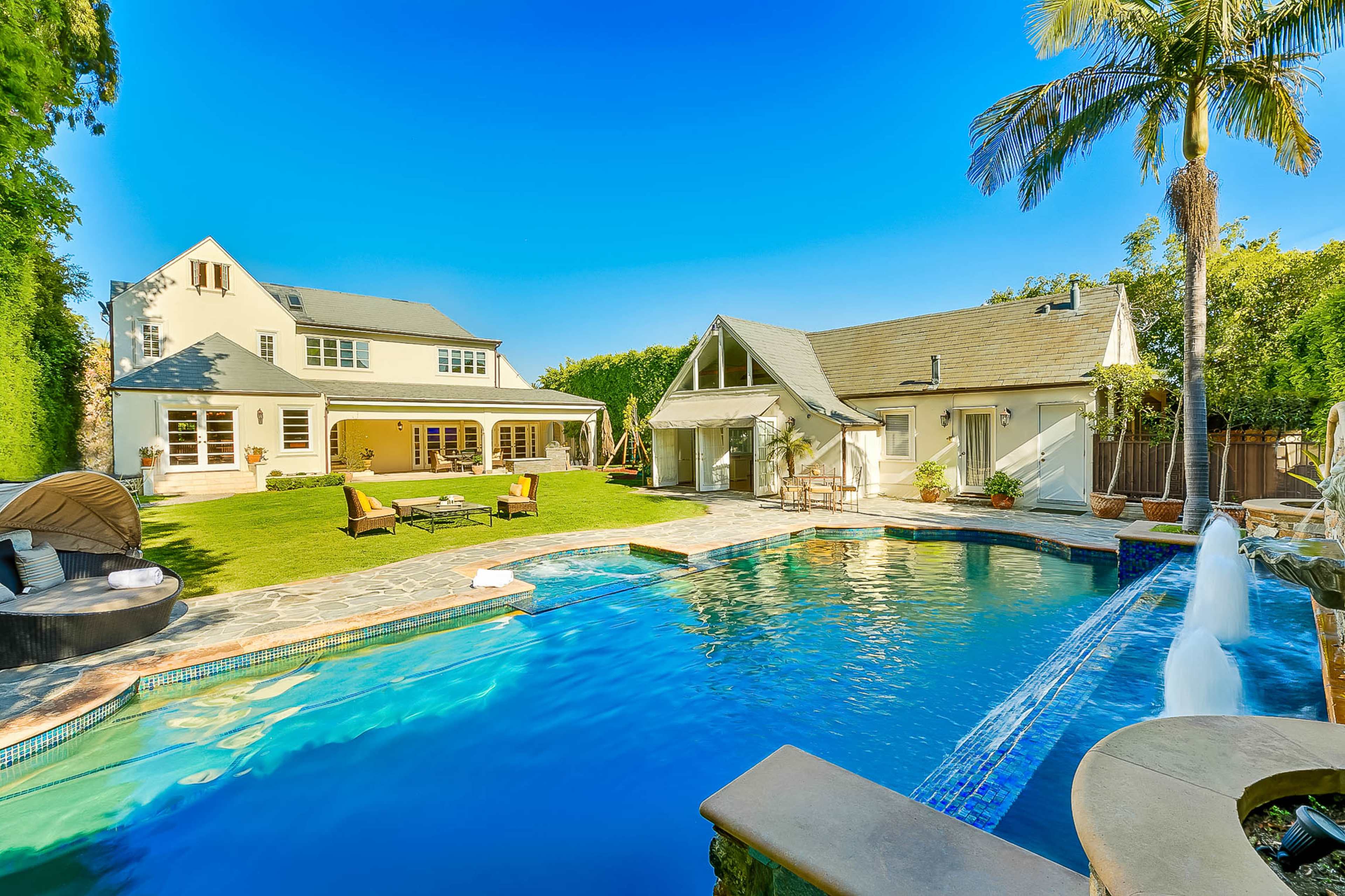 A spacious backyard featuring a swimming pool with a waterfall, surrounded by lounge chairs, a lawn, and two houses in bright sunlight.