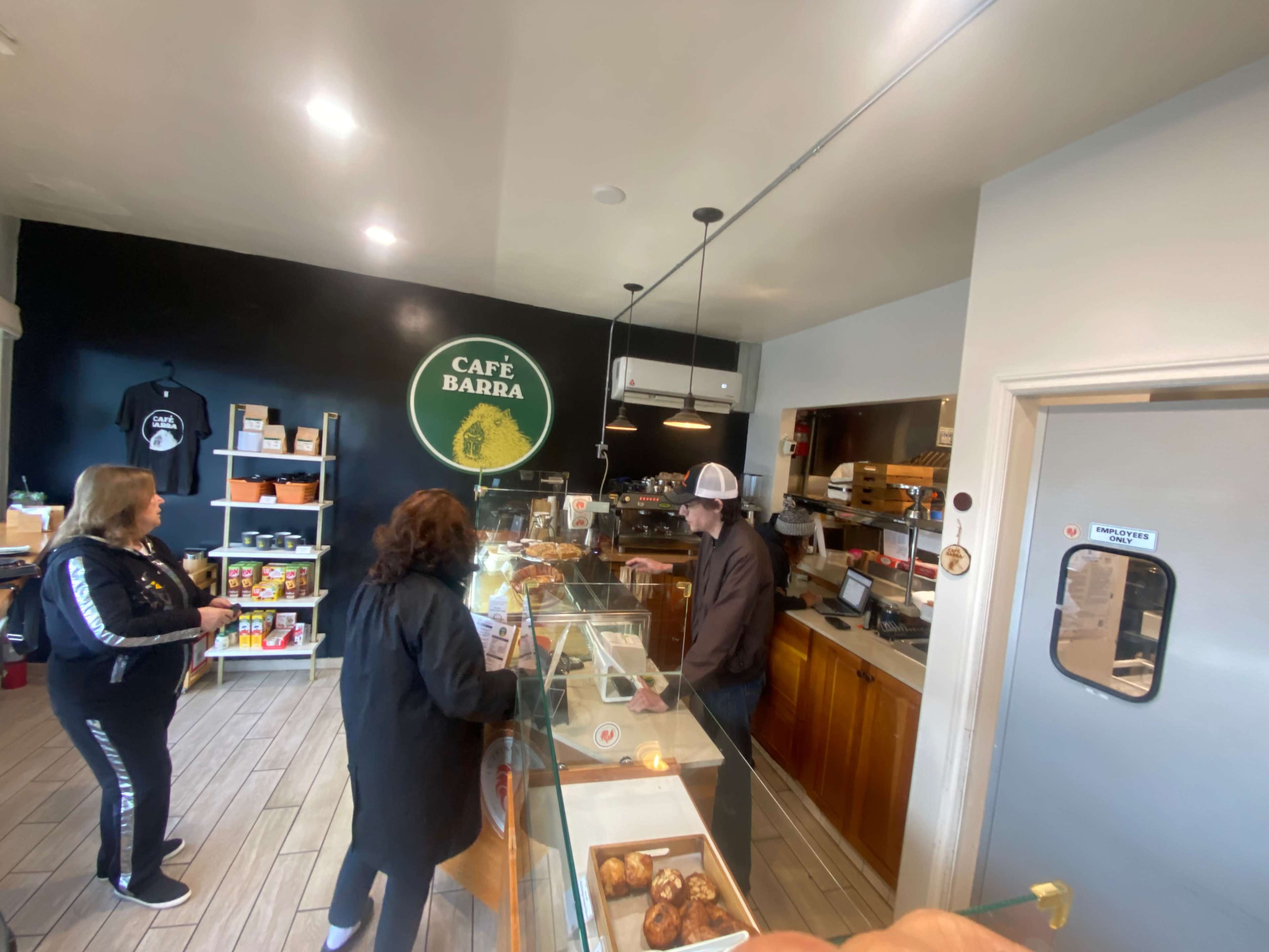 A small café interior shows two customers at the counter while a barista prepares orders behind the glass display.