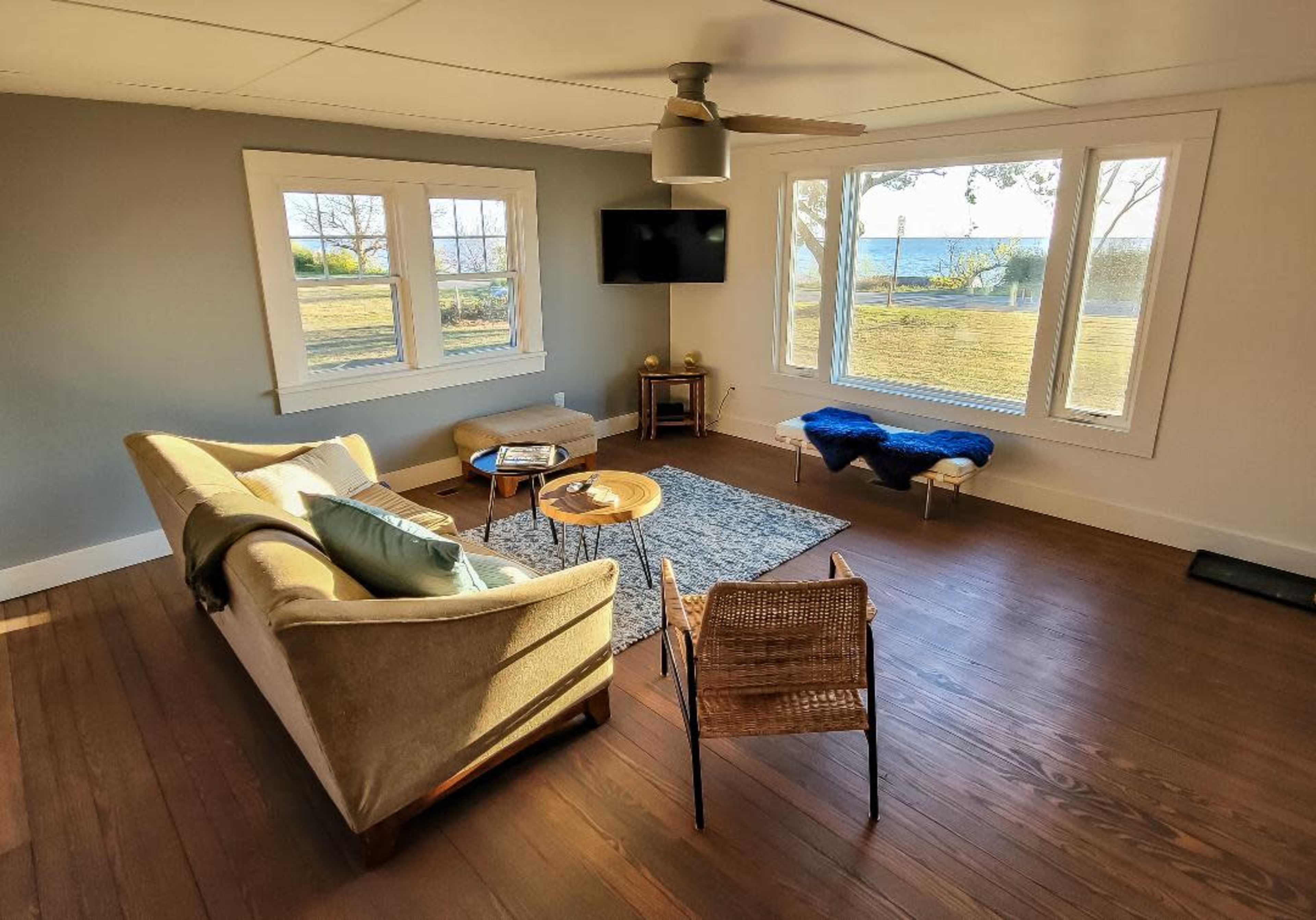 The room features a sofa and a chair arranged around a small coffee table, with large windows showing a view of a grassy area and water beyond.