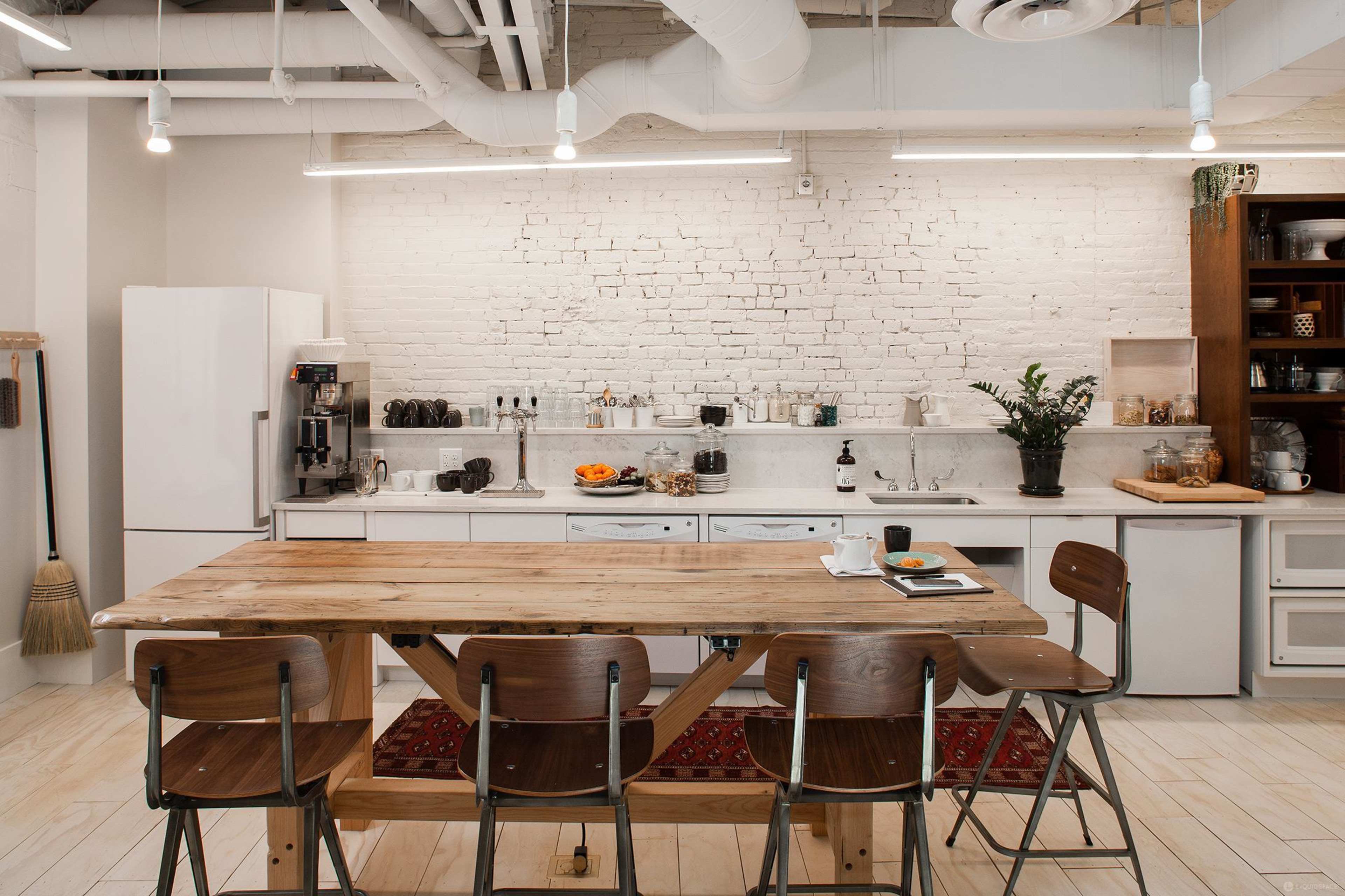 A modern kitchen with a wooden dining table, metal chairs, and a white brick wall featuring kitchen appliances and glassware.
