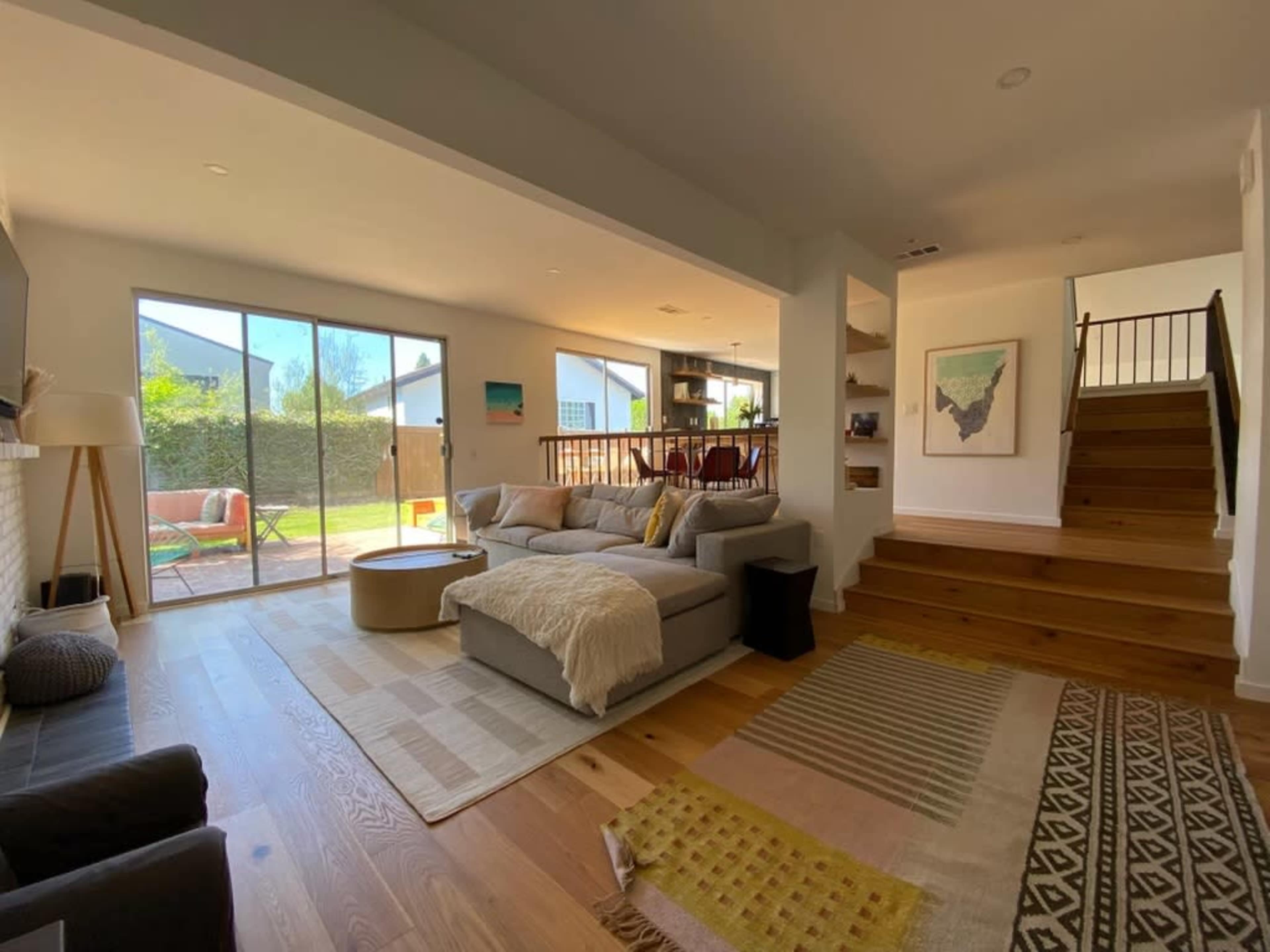 A modern living room with a large sectional sofa, a coffee table, and large windows overlooking a backyard, with a staircase leading to an upper level.
