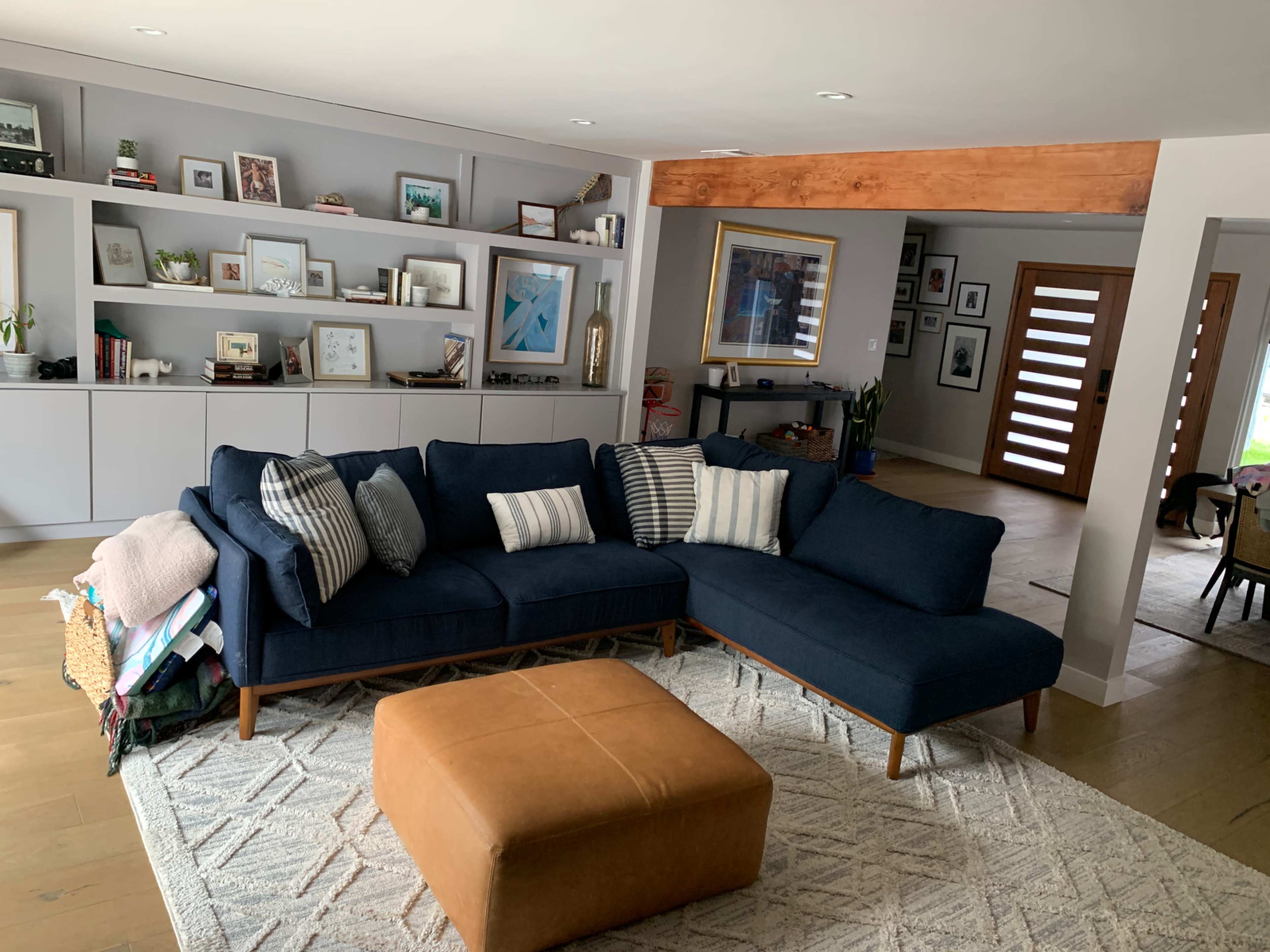A modern living room featuring a navy blue sectional sofa, a large tan ottoman, and a bookshelf with various framed photos and decorative items against a light gray wall.