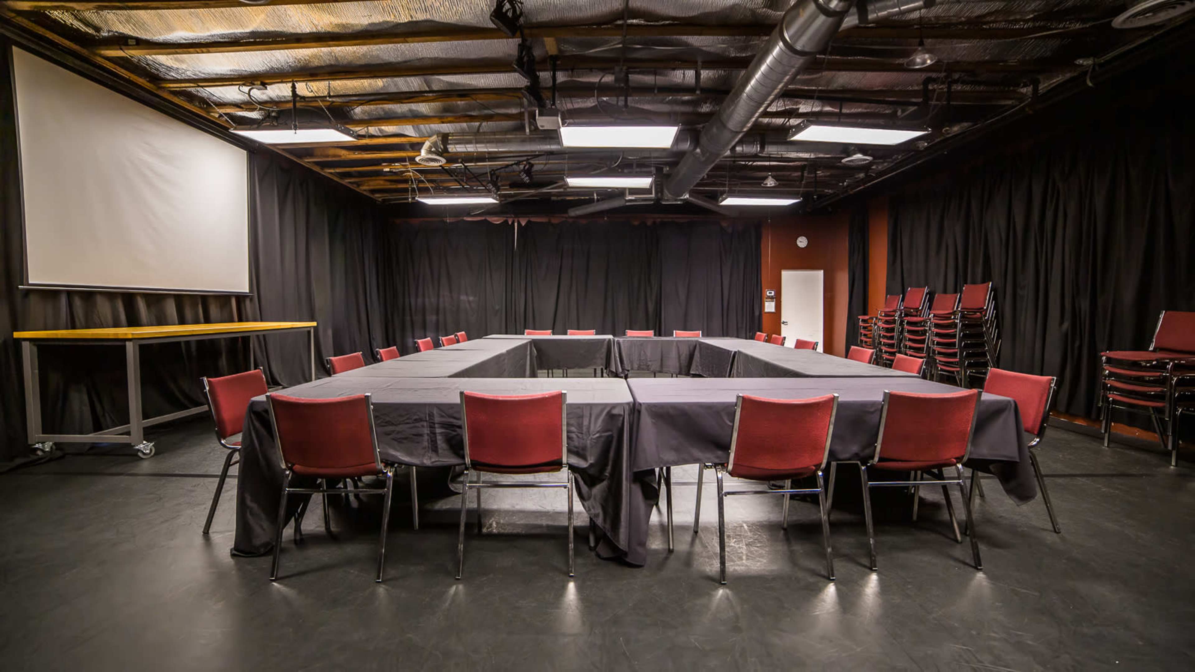 A large meeting room features a square table surrounded by chairs, with a projector screen and a black curtain backdrop.