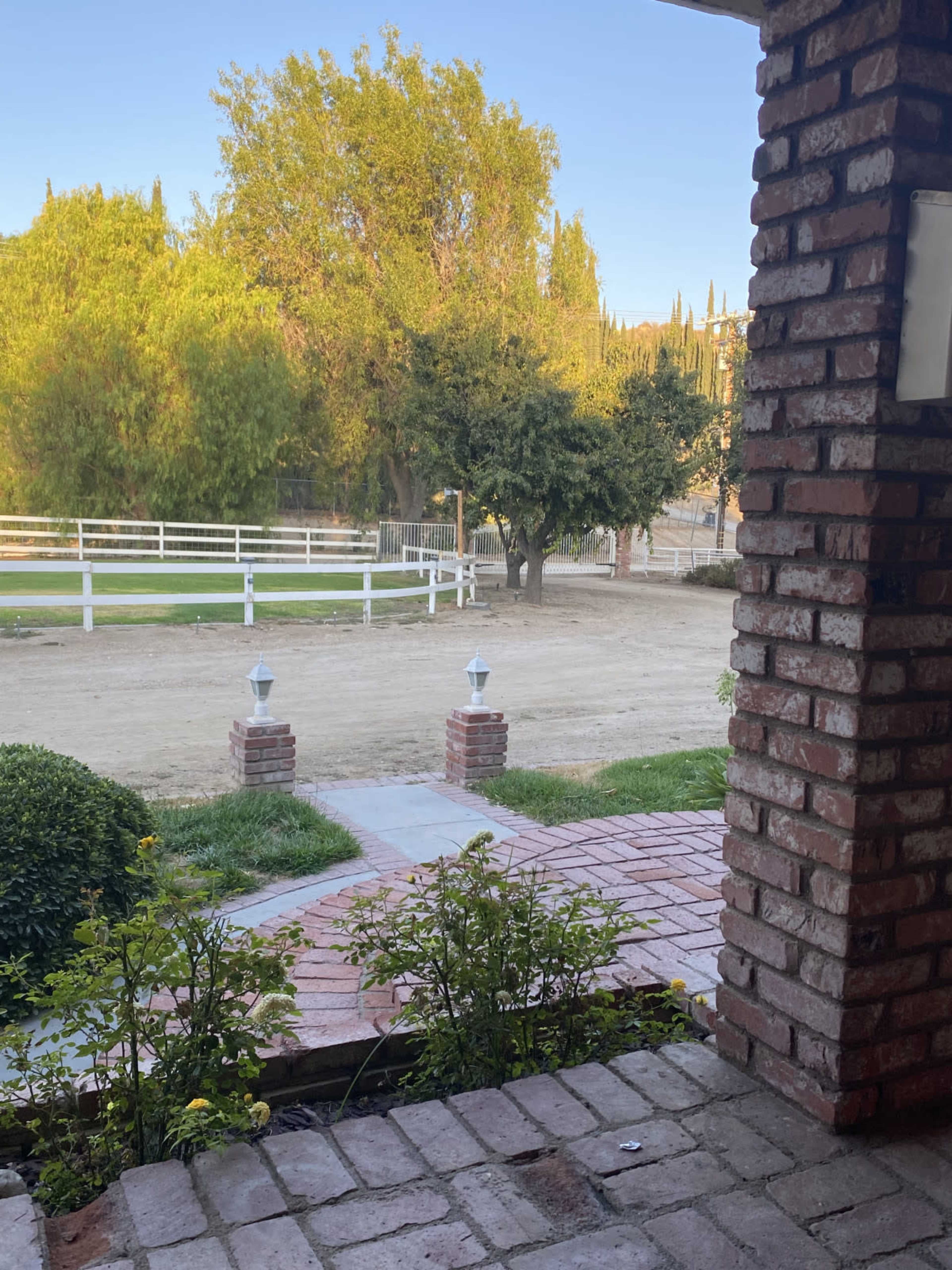 The image shows a view from a brick porch overlooking a dirt path, surrounded by greenery and a white fence in the background.