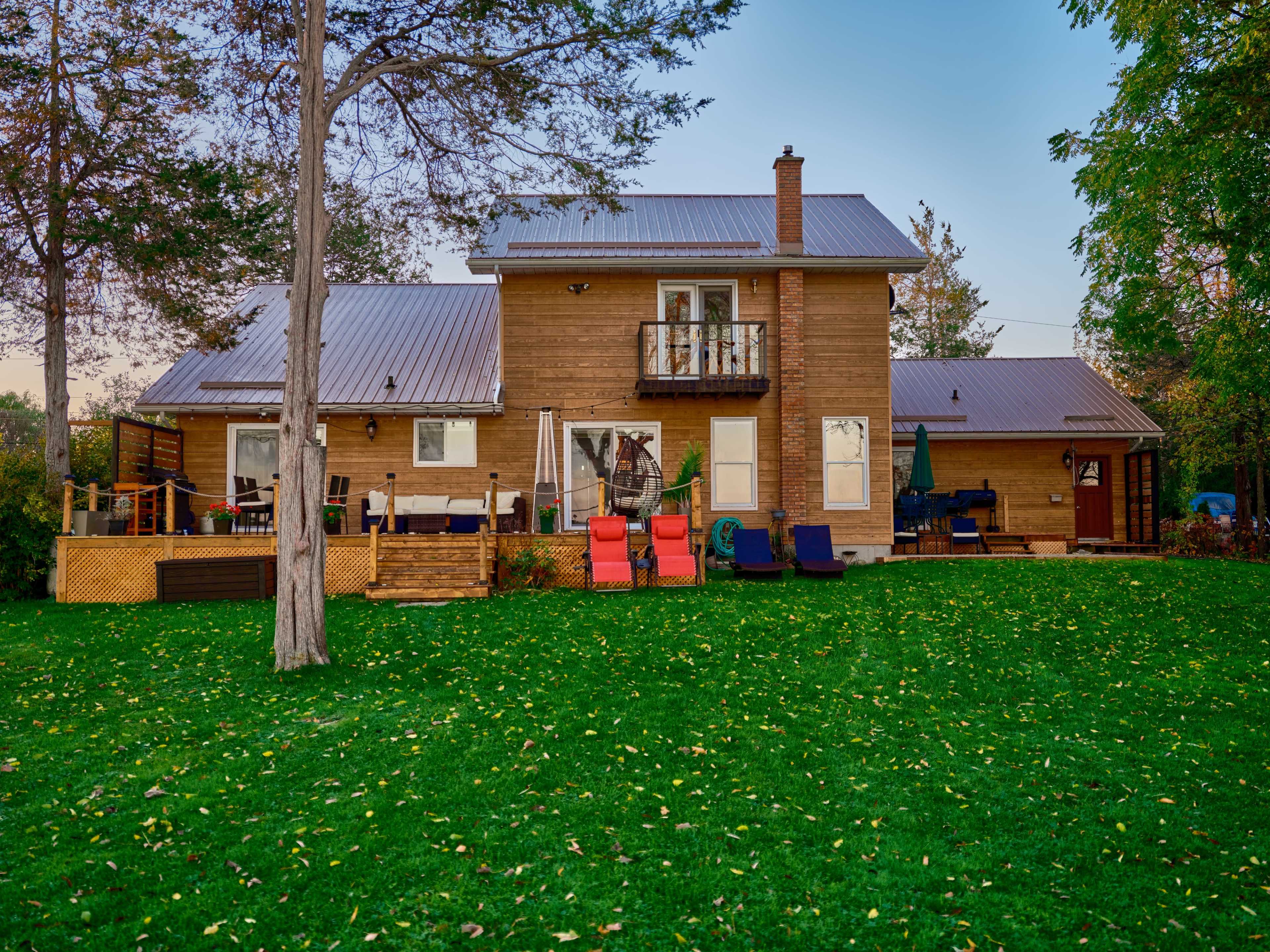 A two-story wooden house with a metal roof is situated in a grassy area surrounded by trees.