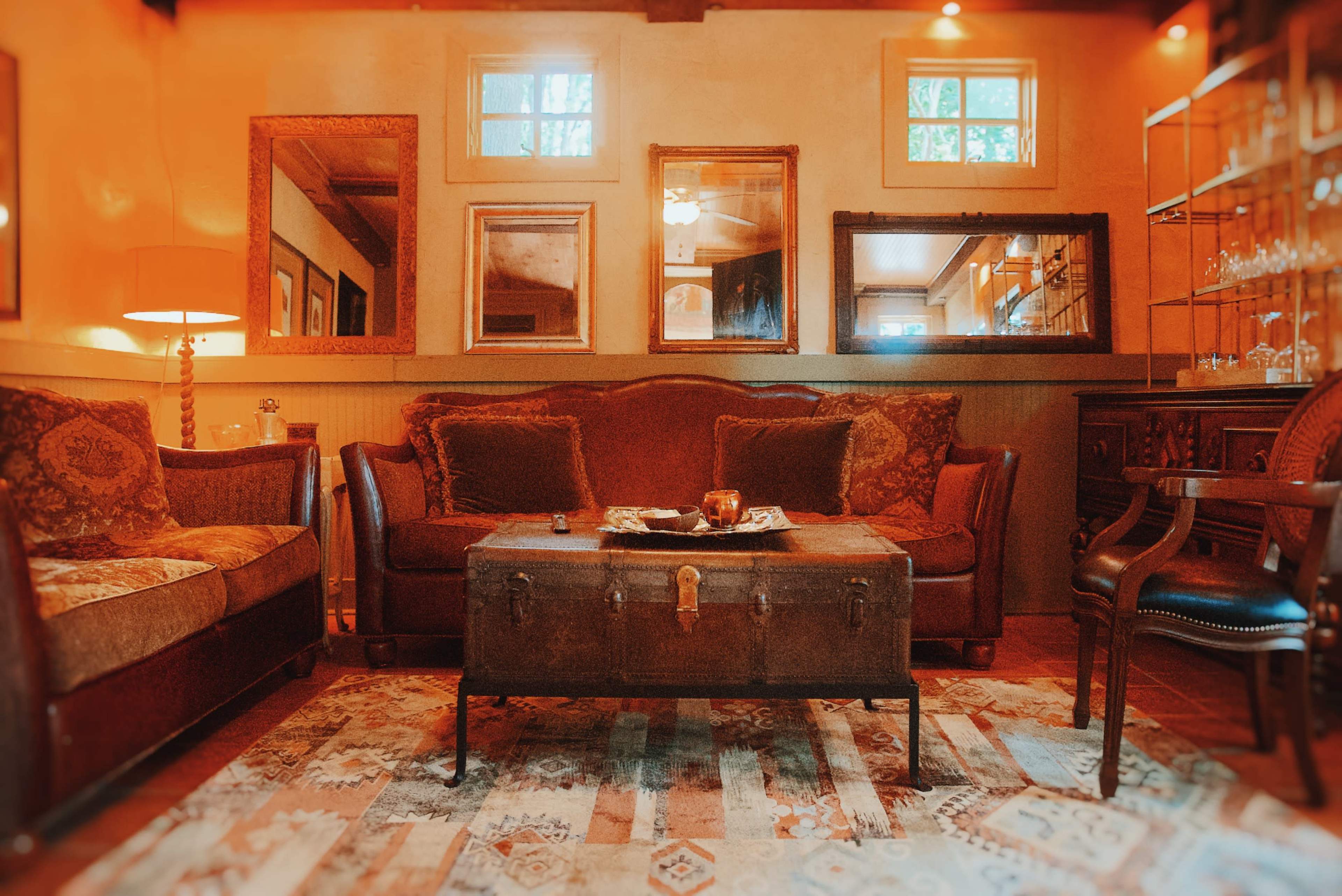 The image depicts a cozy living room with a dark wooden coffee table at the center, surrounded by two brown sofas and decorated with various mirrors and a rug.