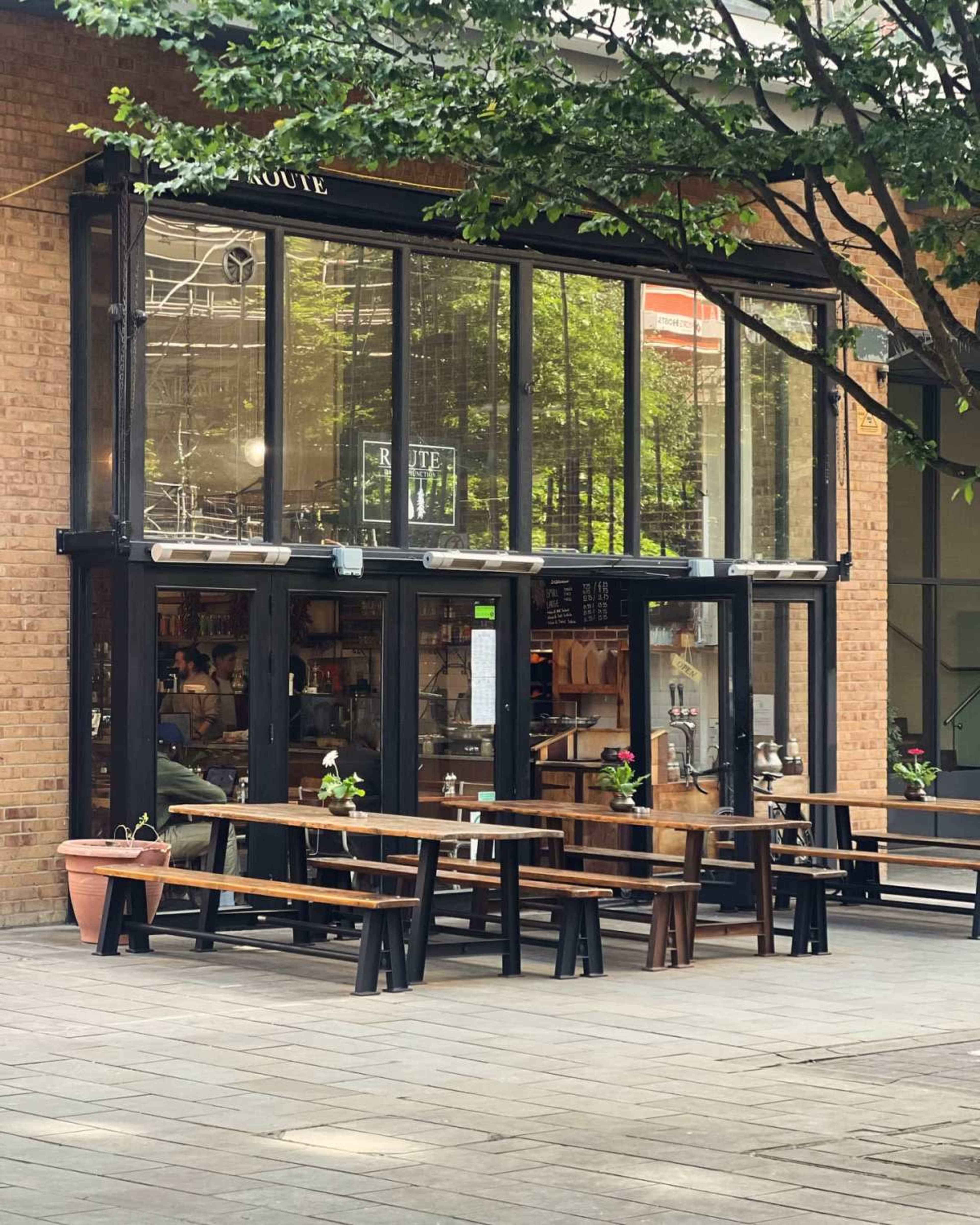A café with large glass windows features outdoor seating made of wooden benches and tables set on a stone pathway.