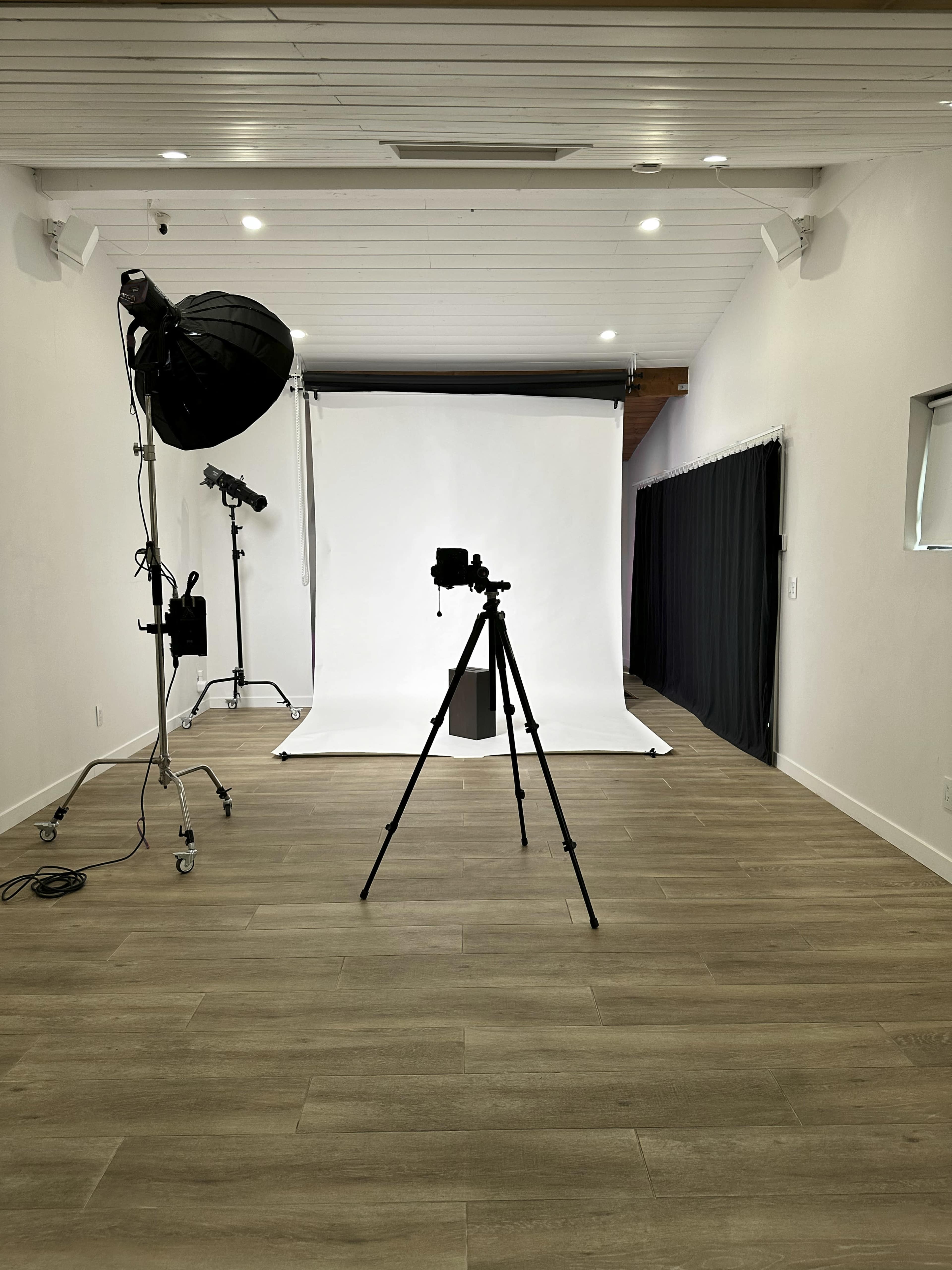 An empty photography studio features a tripod and camera facing a white backdrop, with a softbox and other lighting equipment positioned nearby.