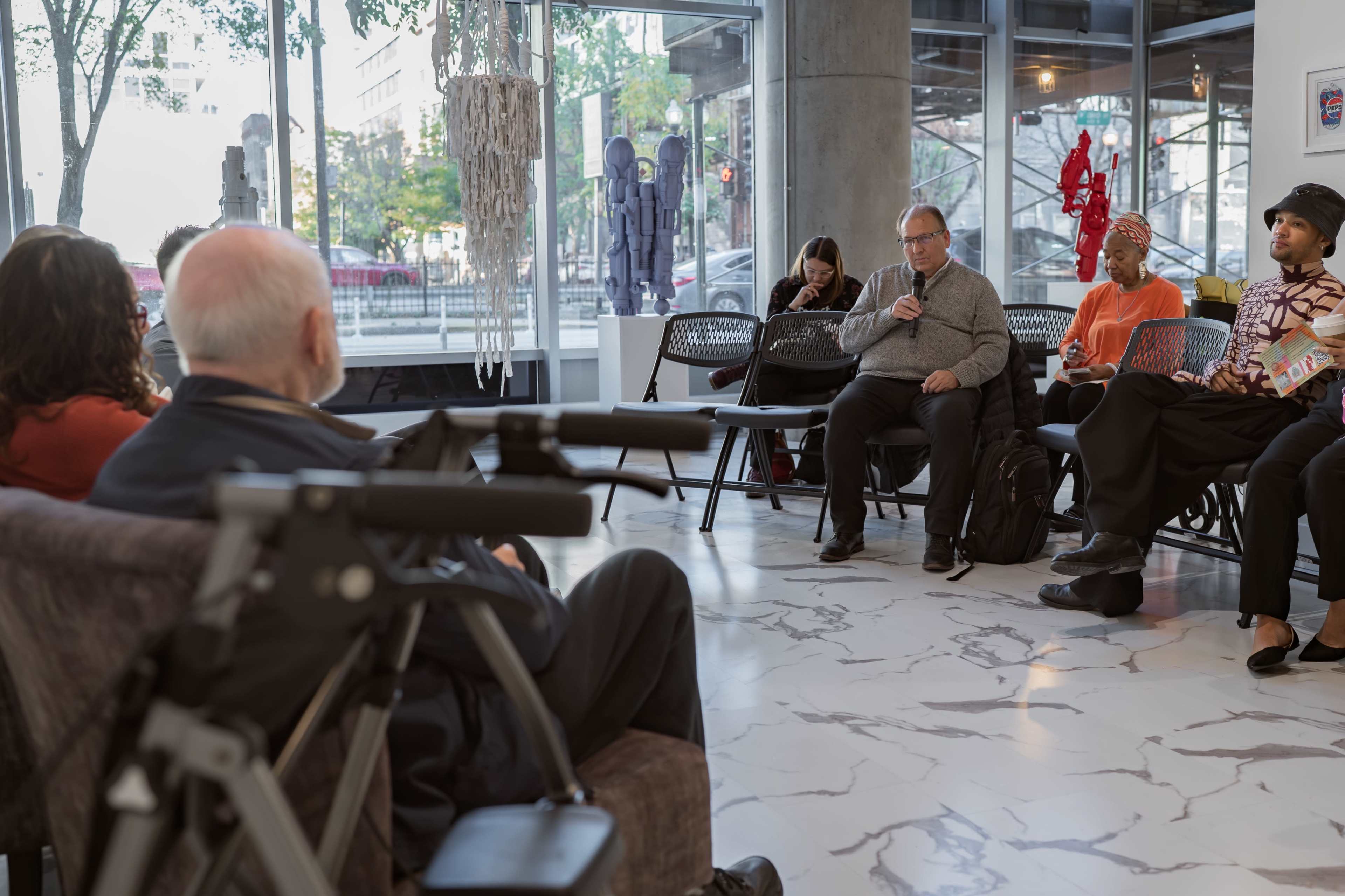 A group of people is seated in a modern, well-lit space with large windows, some engaged in conversation while others are focused on their phones.