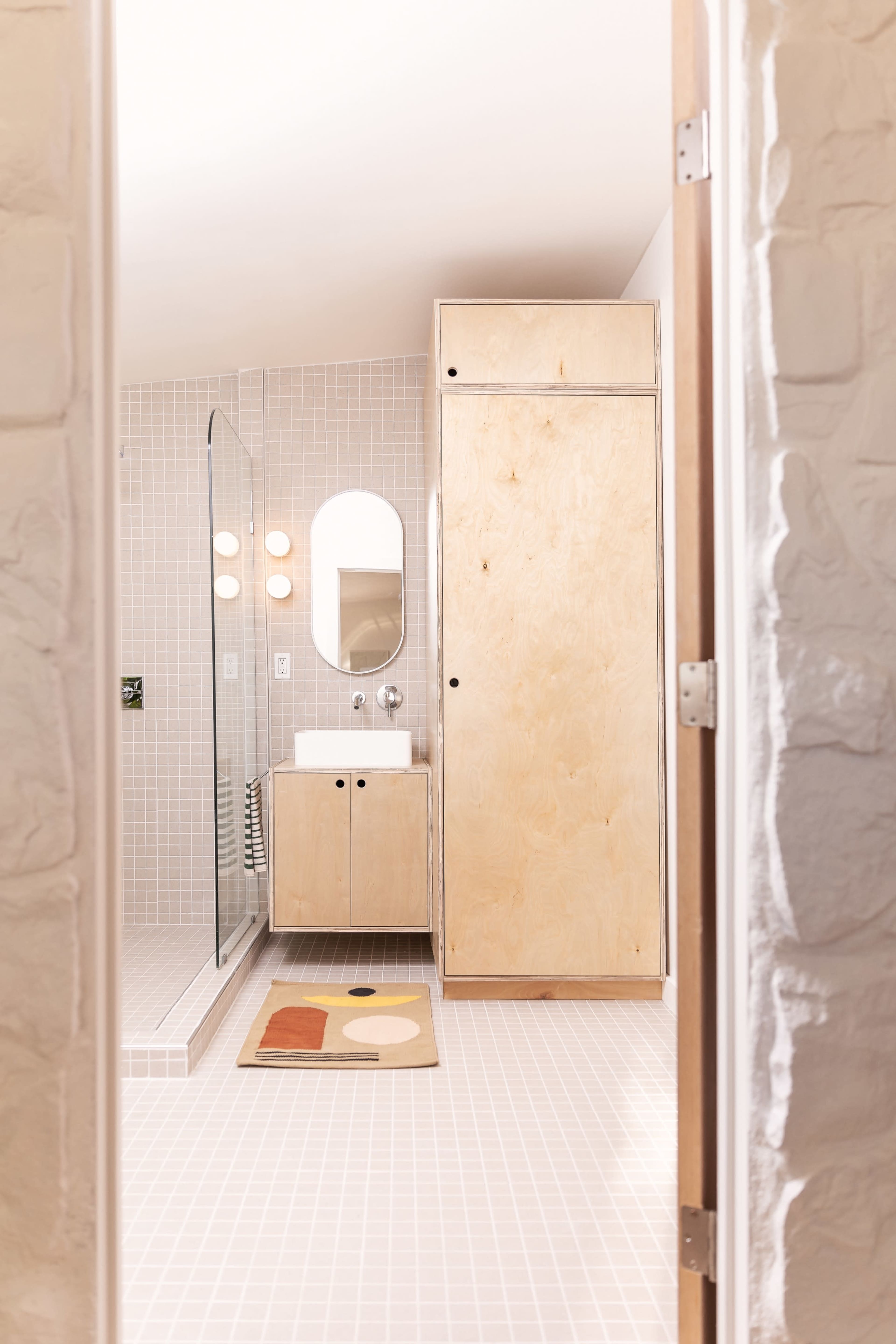 A modern bathroom with a wooden cabinet, a round mirror, and a glass shower enclosed by tiled walls and floor.