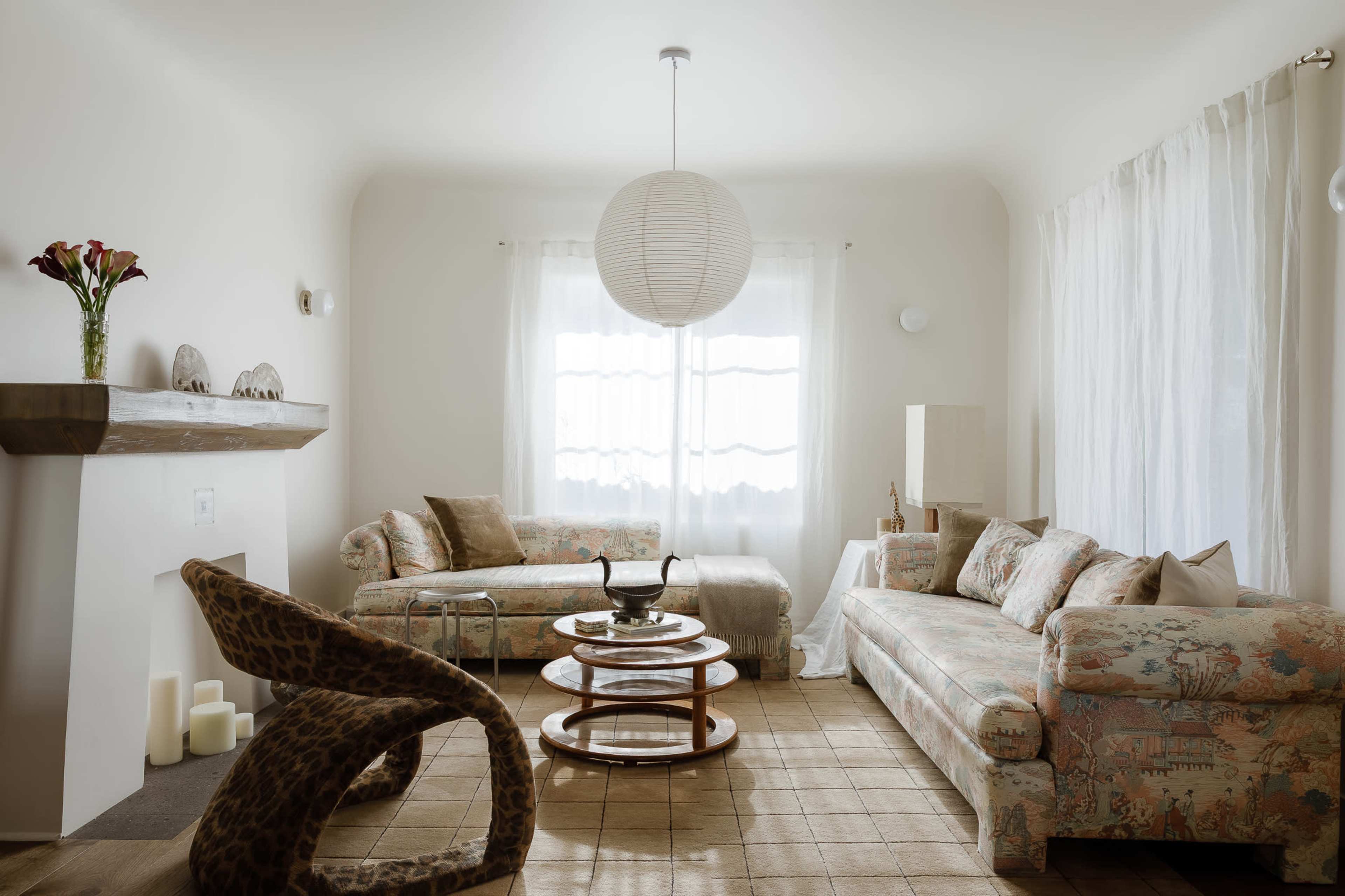 A bright living room features two floral-patterned sofas, a round coffee table, and a large paper lantern hanging from the ceiling, with sunlight streaming through sheer curtains.