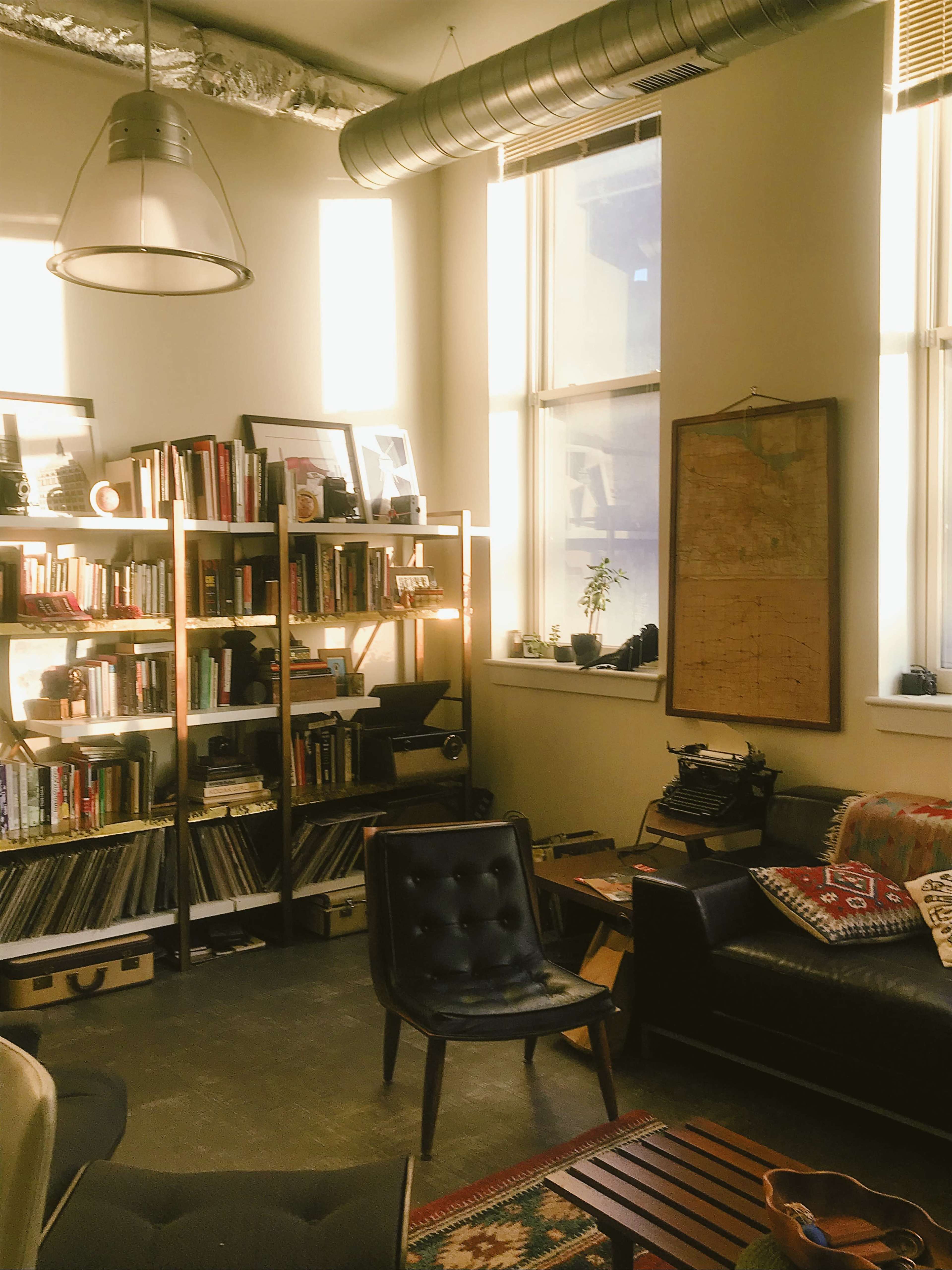A cozy reading nook featuring a bookshelf filled with books, a vintage typewriter, and a comfortable seating area illuminated by natural light from large windows.