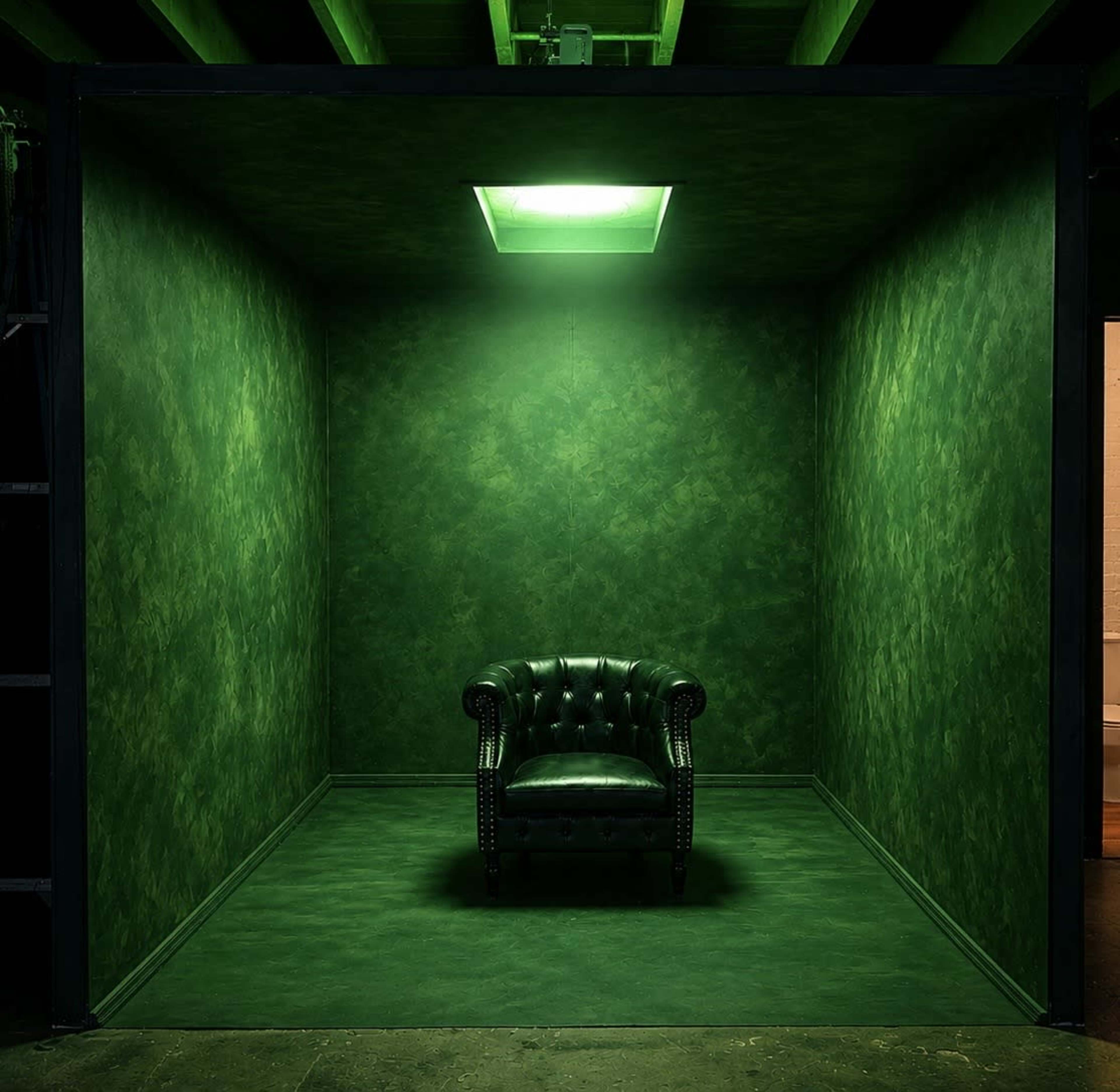 A solitary green armchair sits in a small, dimly lit room with green walls and a skylight.