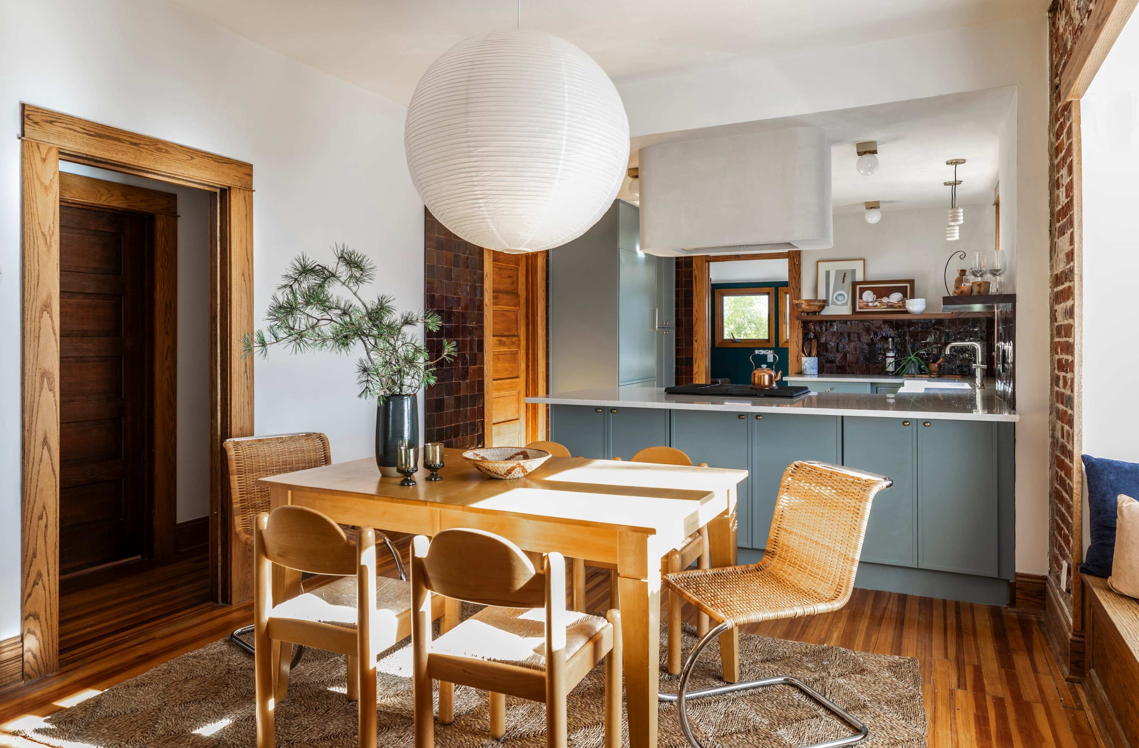 A dining area with a wooden table and chairs, a round paper lantern hanging overhead, and a kitchen area visible in the background.