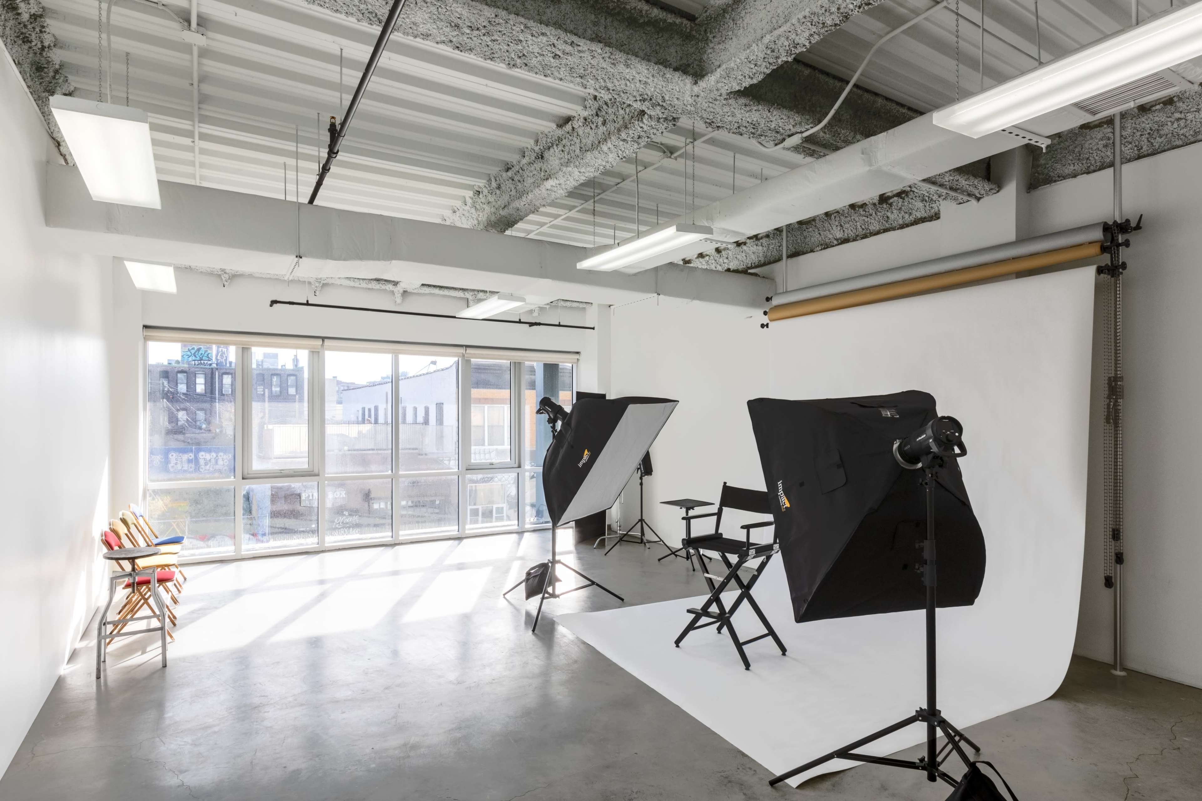 A photography studio equipped with two lighting setups, a backdrop, and a director's chair near large windows that allow natural light.