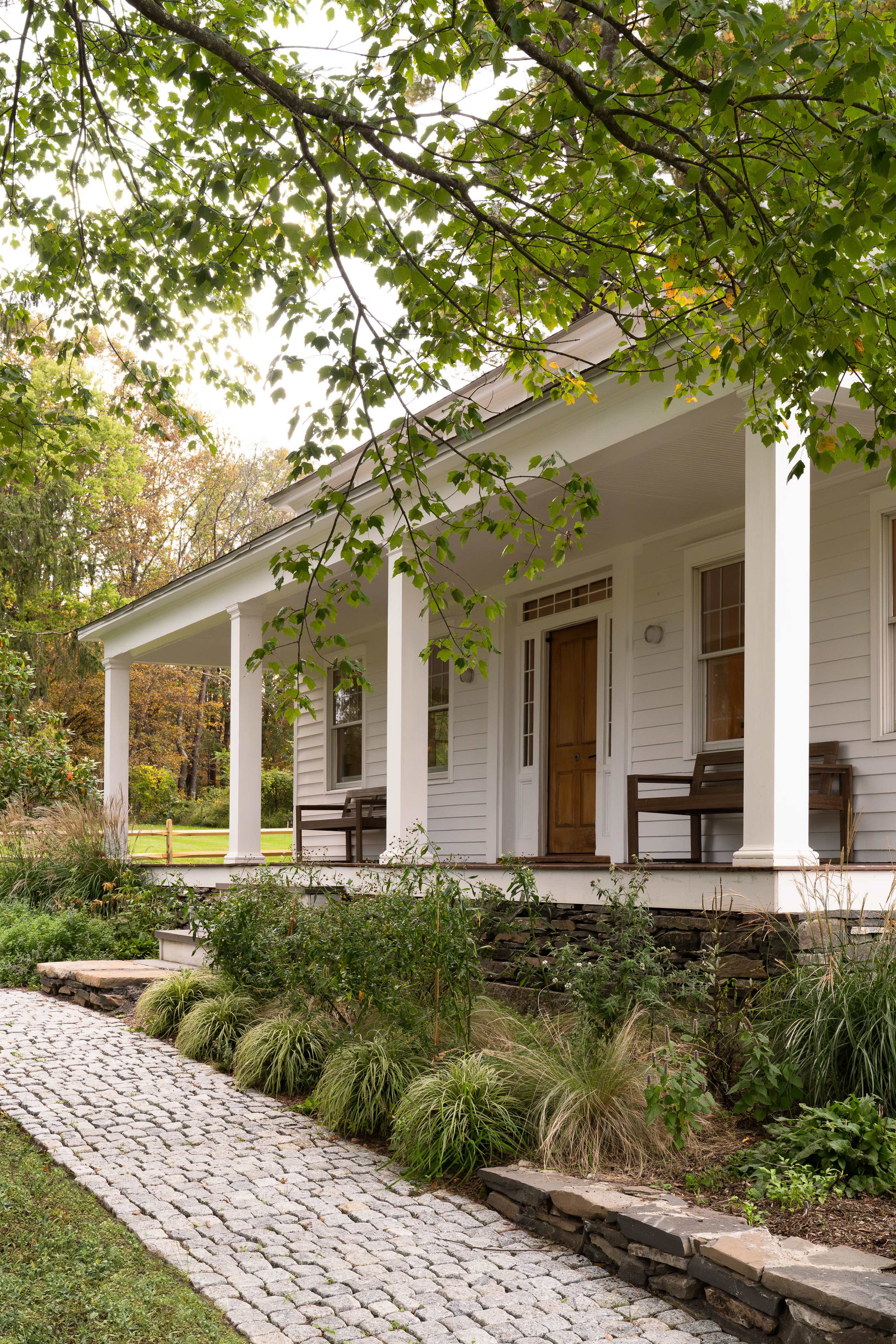 A white house with a front porch is flanked by two benches and surrounded by landscaped greenery, leading to a stone pathway.